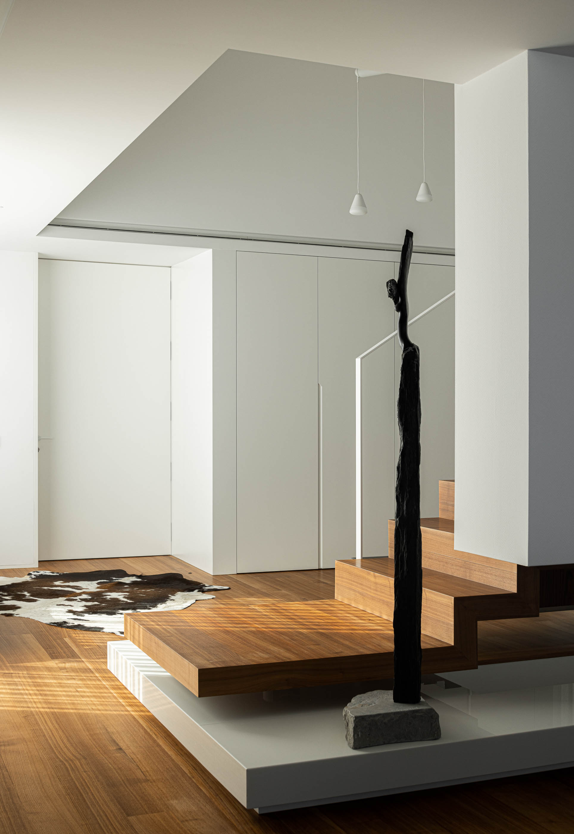 Hall de entrada: Escada flutuante em madeira, alcatifa de pele e candeeiros suspensos em espaço minimalista branco. — Riscowhite arquitetura Risco Singular em Barcelos