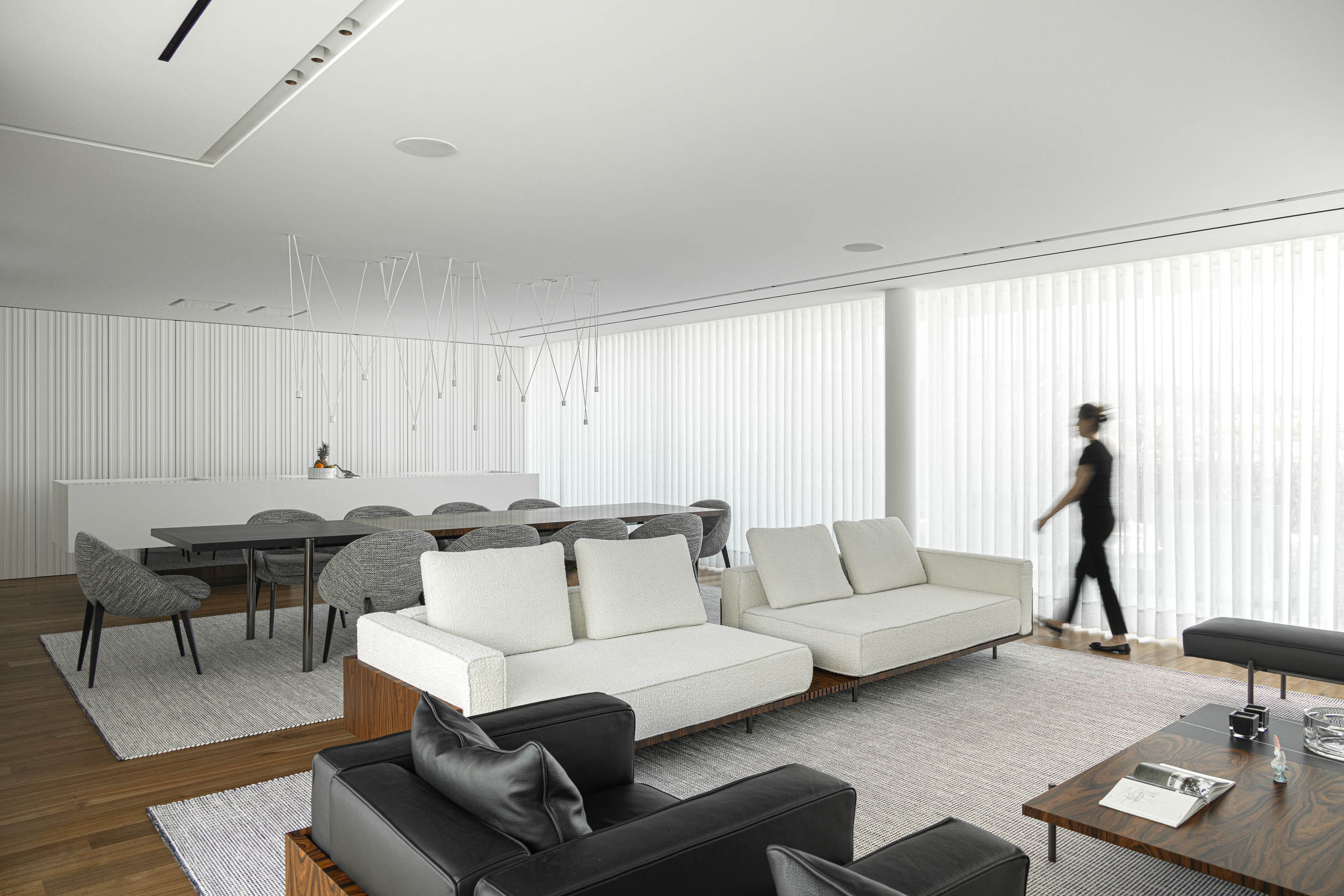 Sala de estar: Vista ampla da sala com sofás, mesa de jantar e cortinas verticais brancas. — Riscowhite arquitetura Risco Singular em Barcelos