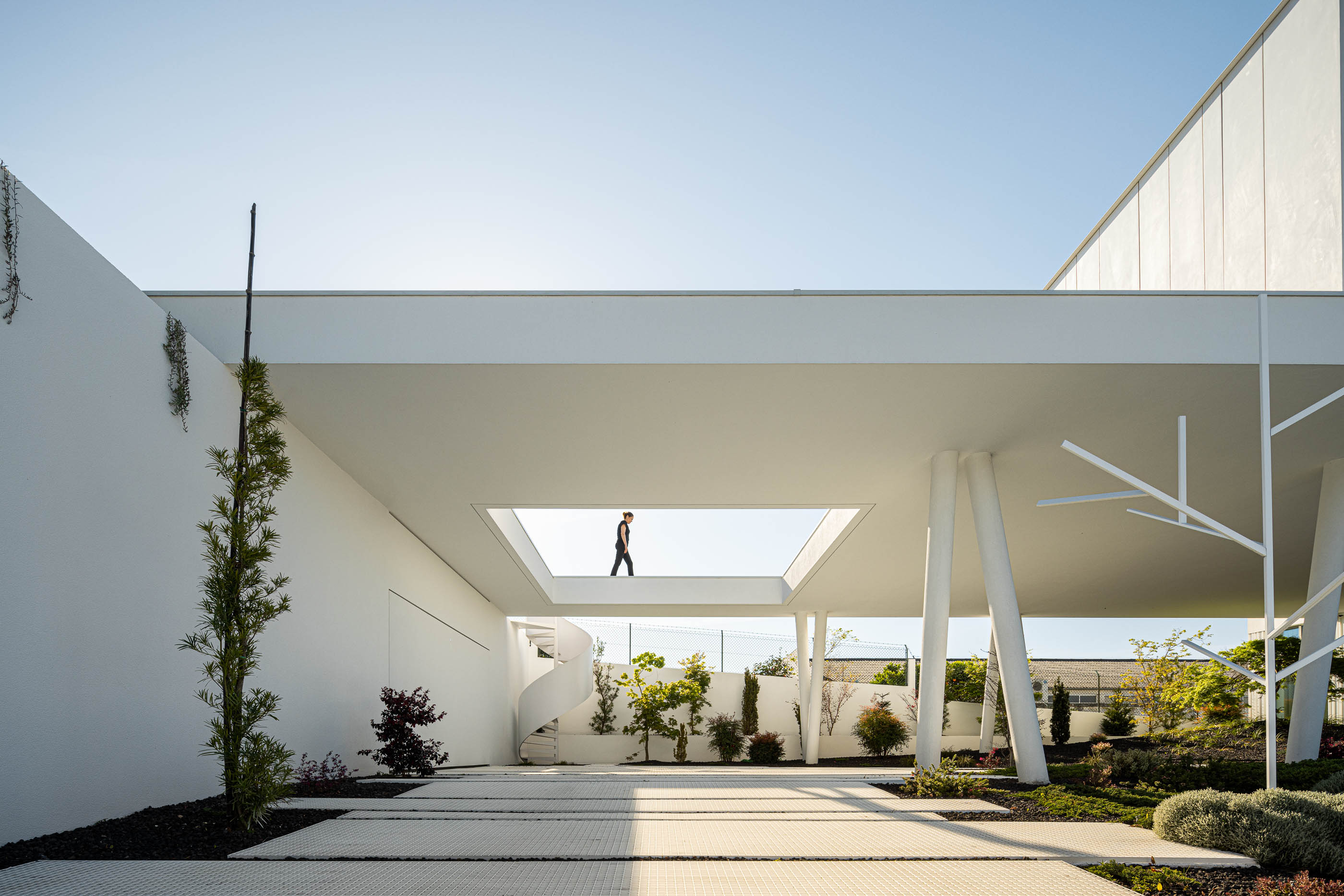 Exterior: Pátio com escada em caracol, jardim minimalista e passadiço elevado com pessoa. — Riscowhite arquitetura Risco Singular em Barcelos