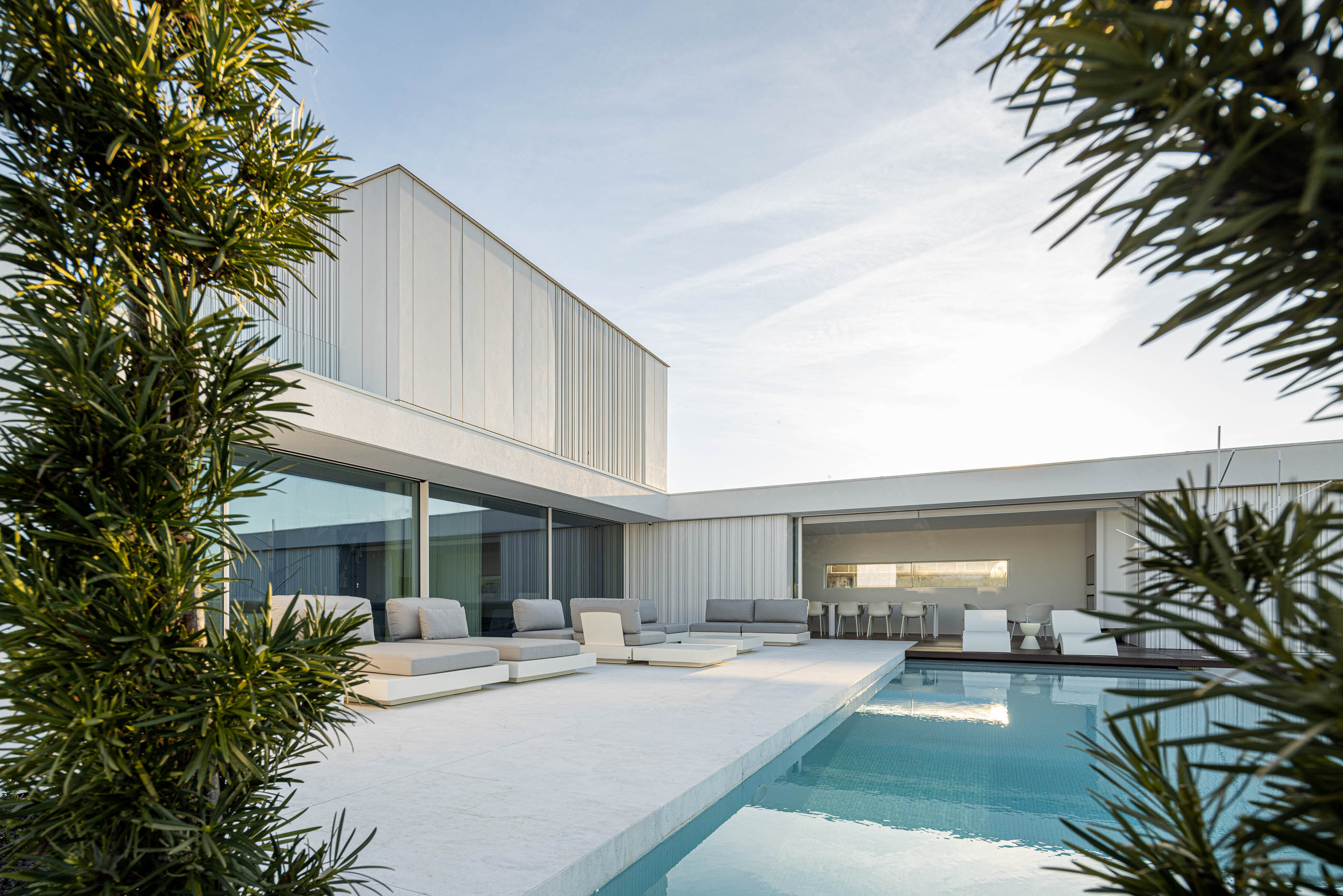 Piscina: Espreguiçadeiras cinzentas junto à piscina com vista para a moradia e zona de refeições exterior. — Riscowhite arquitetura Risco Singular em Barcelos