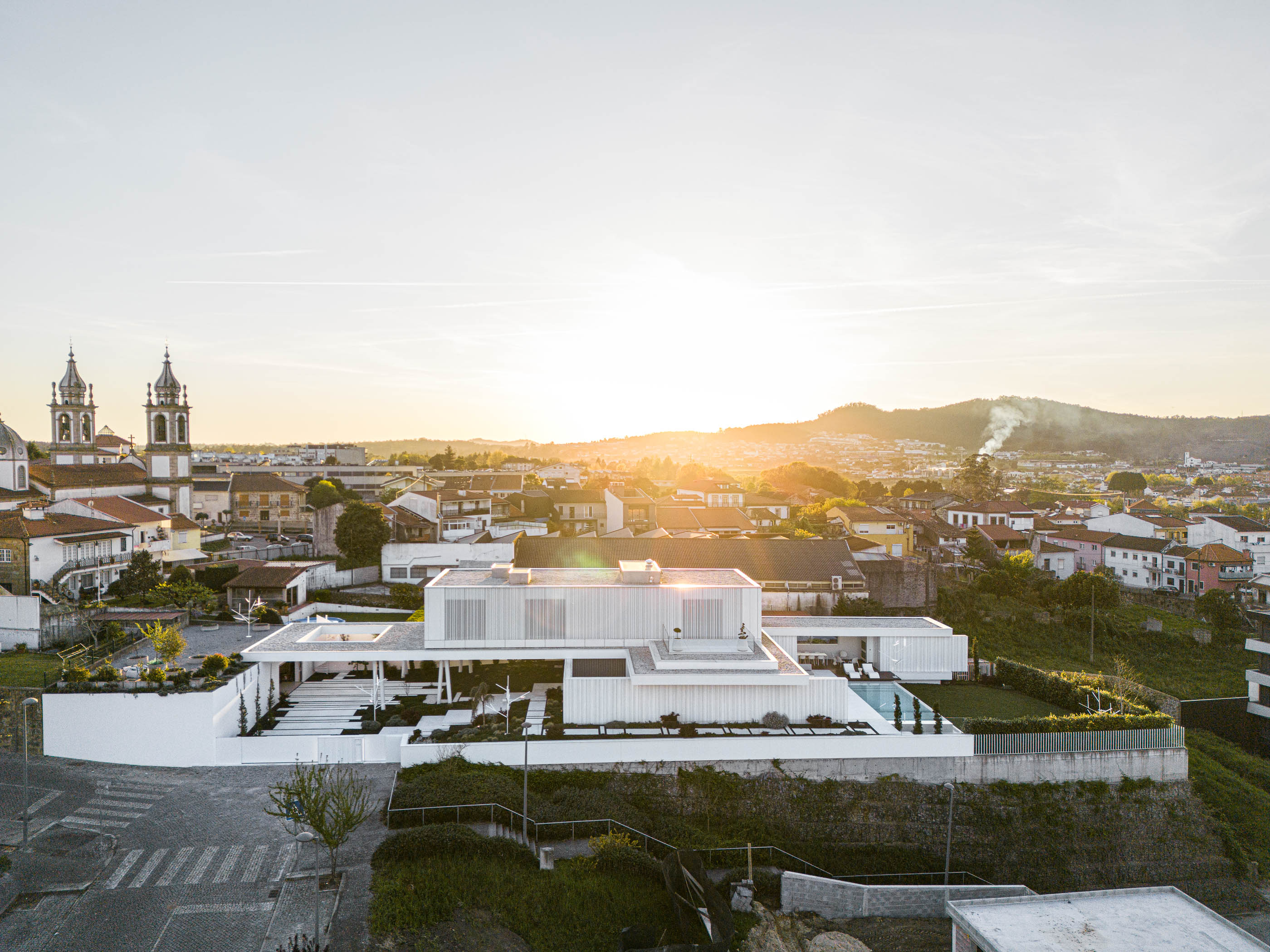 Exterior: Vista panorâmica da moradia branca com piscina, integrada na paisagem de Barcelos ao pôr do sol. — Riscowhite arquitetura Risco Singular em Barcelos