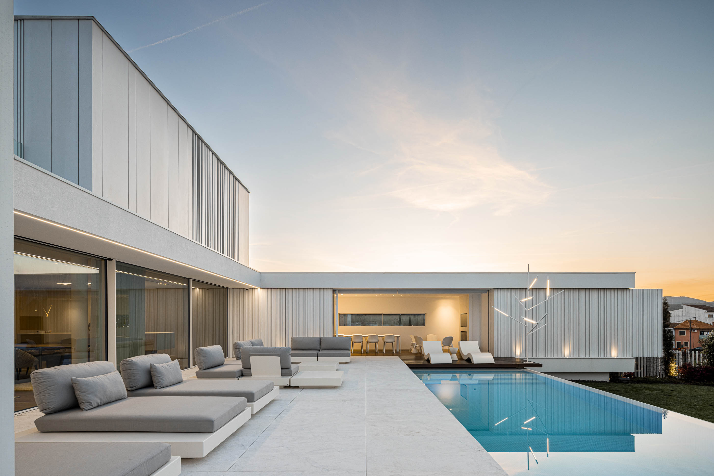 Piscina: Espreguiçadeiras cinzentas junto à piscina com vista para a moradia e zona de refeições exterior. — Riscowhite arquitetura Risco Singular em Barcelos