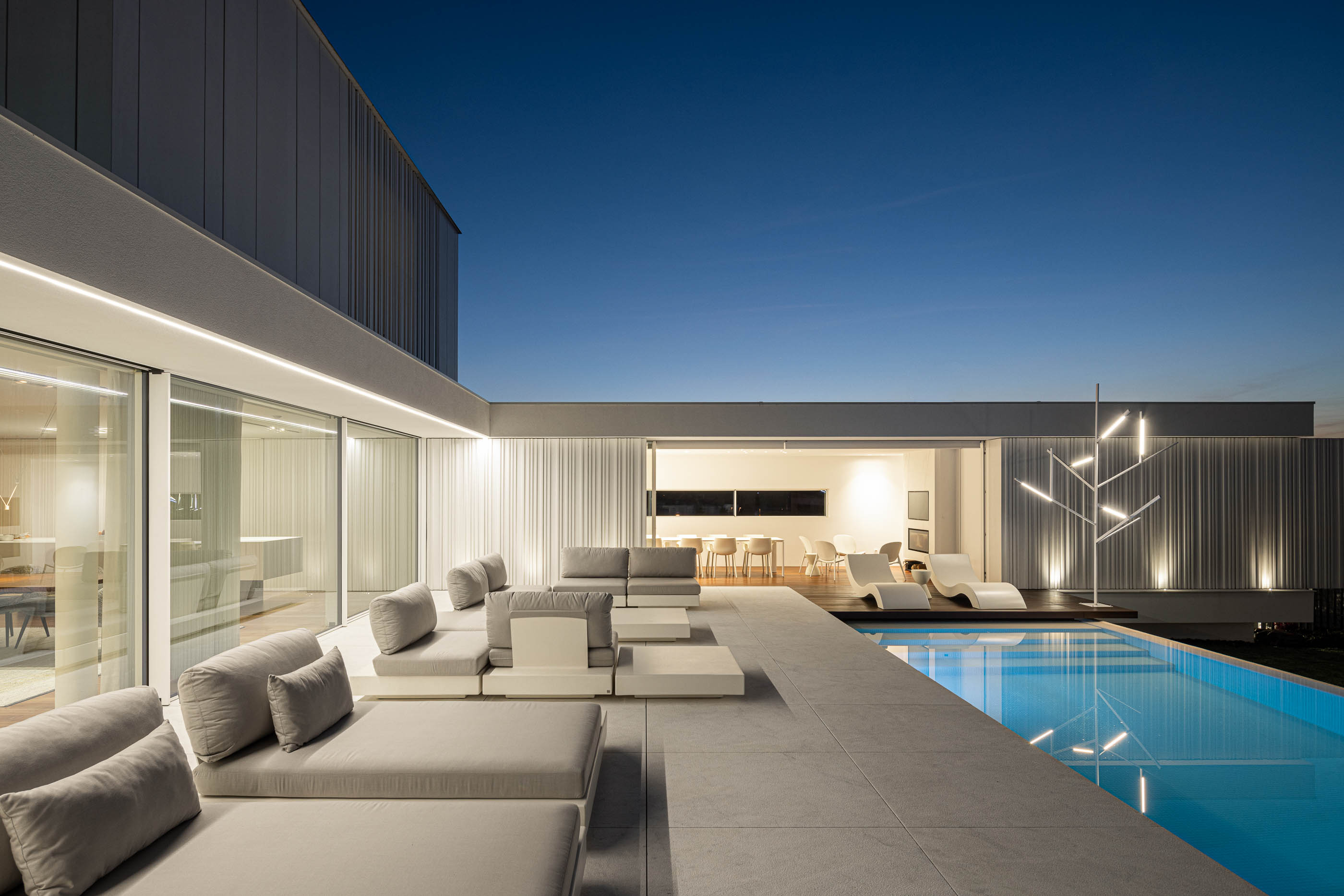 Piscina: Espreguiçadeiras cinzentas junto à piscina com vista para a moradia iluminada à noite. — Riscowhite arquitetura Risco Singular em Barcelos