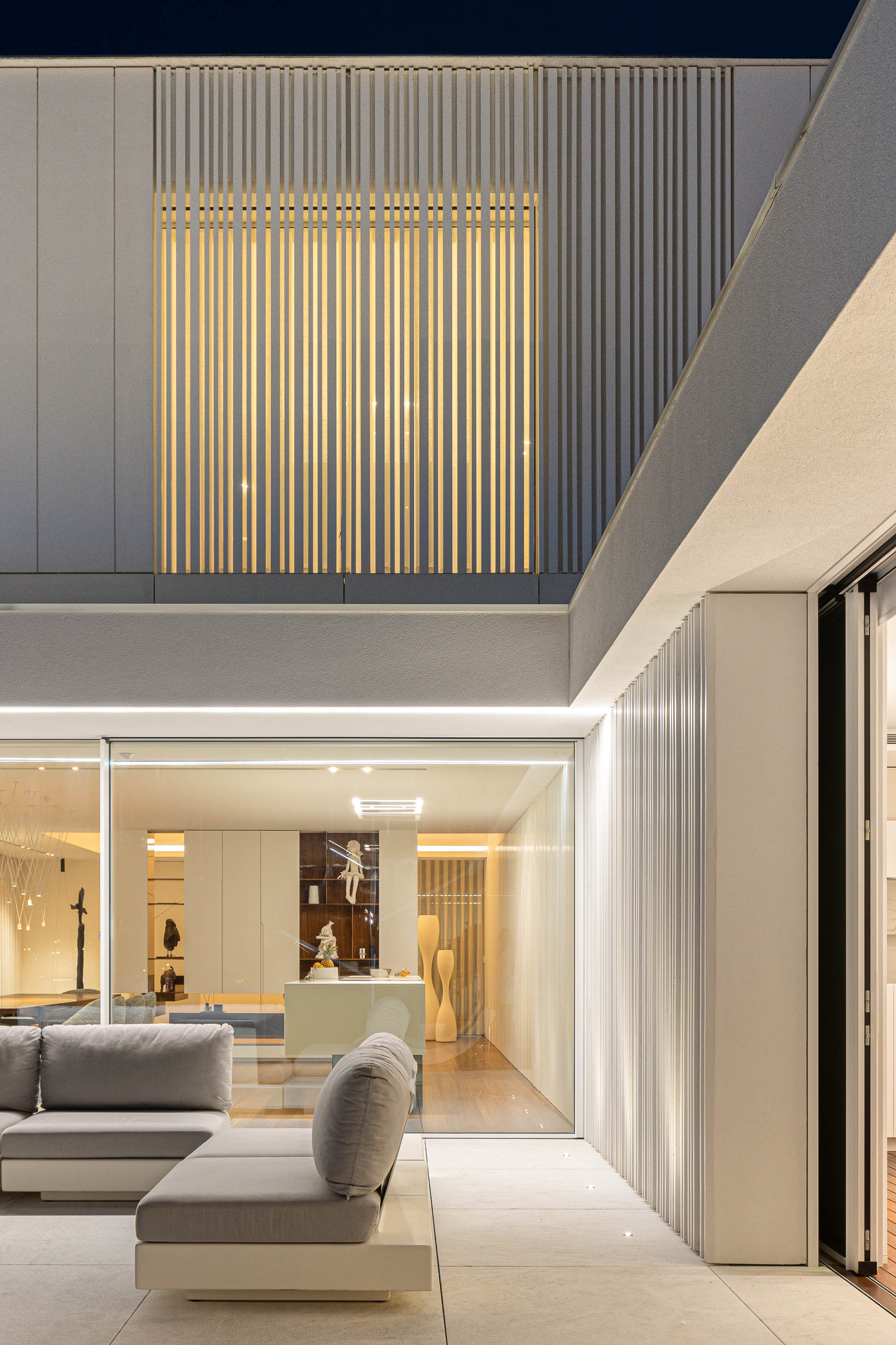 Exterior: Detalhe da fachada iluminada, com zona de estar exterior e vista para o interior da moradia. — Riscowhite arquitetura Risco Singular em Barcelos