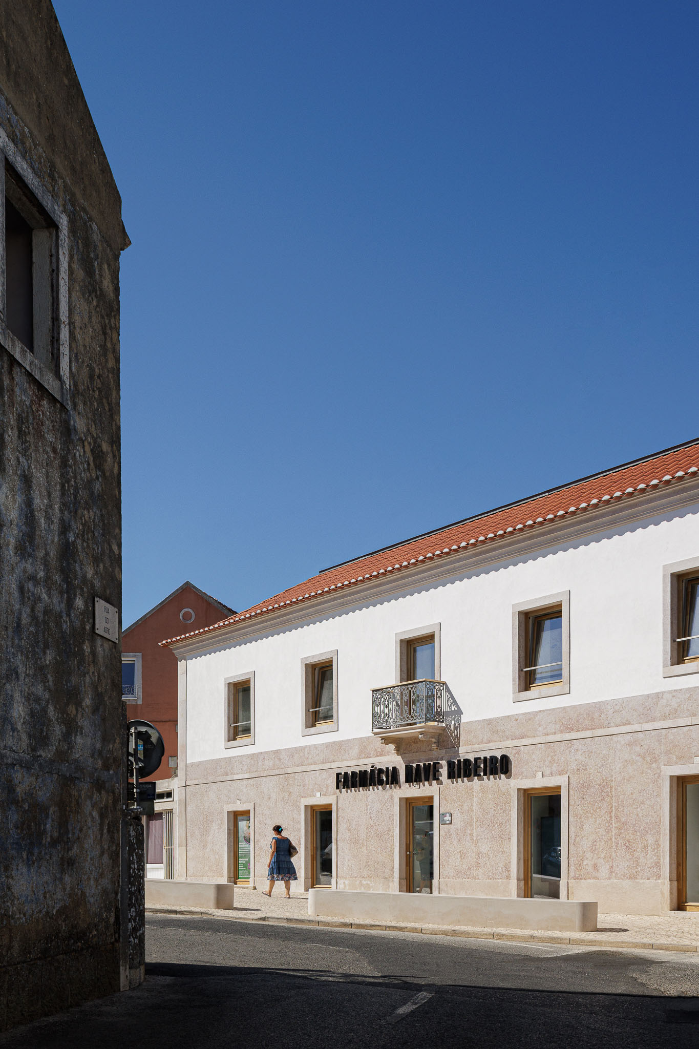 Fachada: Edifício de dois pisos com fachada em pedra e reboco branco, telhado cerâmico e varanda em ferro forjado. — Farmácia NR arquitetura Esquissos Arquitectura em Sintra