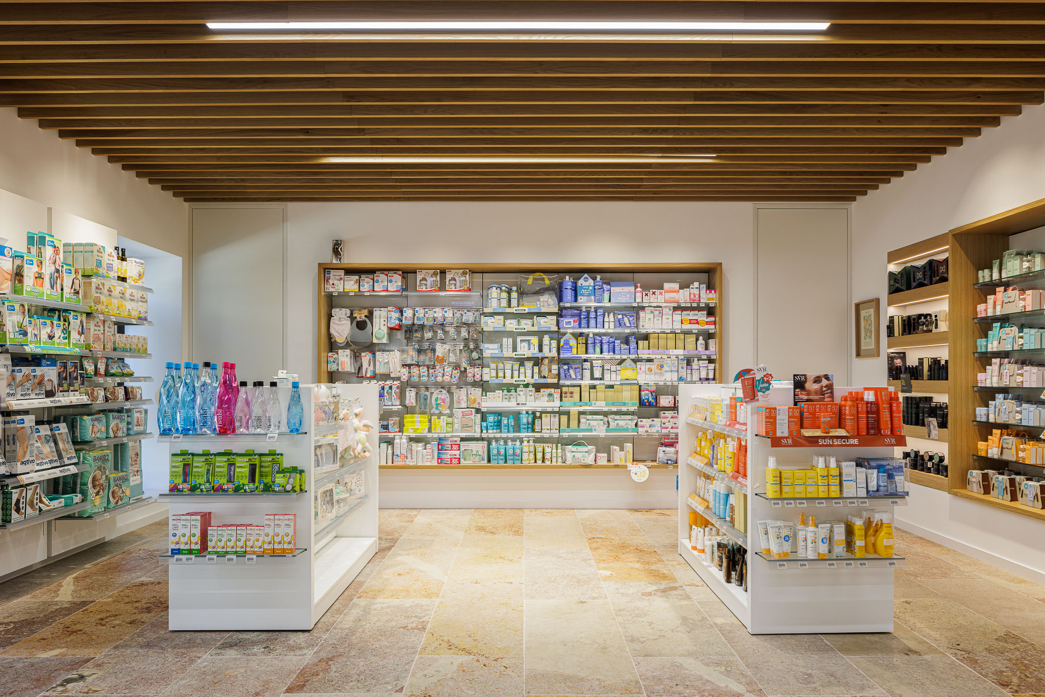 Interior: Vista geral da área de vendas com prateleiras repletas de produtos, pavimento em pedra e teto em madeira. — Farmácia NR arquitetura Esquissos Arquitectura em Sintra