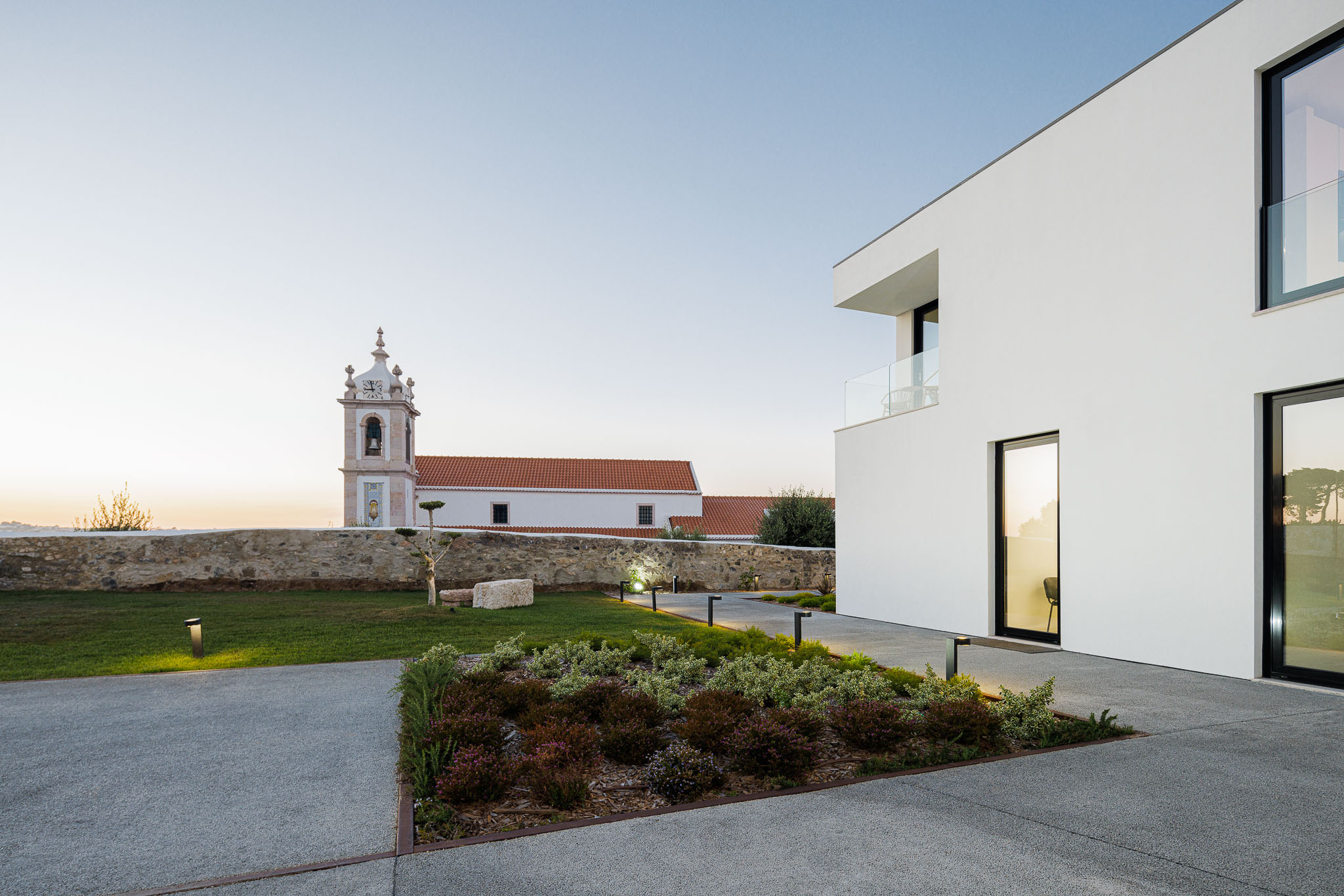 Exterior: Edifício moderno com jardim relvado, iluminação exterior e vista para a igreja ao fundo. — Farmácia NR arquitetura Esquissos Arquitectura em Sintra