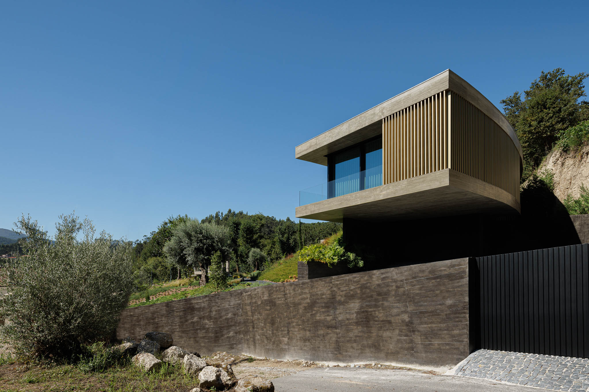 Exterior: Vista da casa moderna em betão e ripas douradas, suspensa sobre um muro de pedra rústico. — Casa de Bouro arquitetura Mutant Architecture and Design em Terras de Bouro
