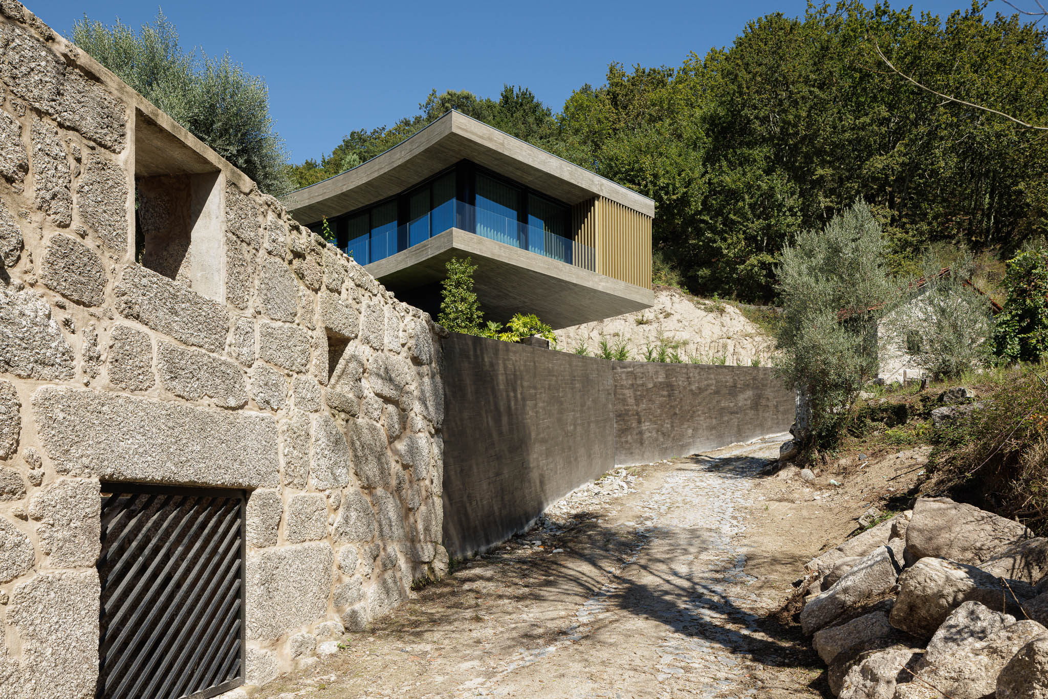 Exterior: Acesso à casa por caminho de pedra, com muro de betão e construção antiga em pedra à esquerda. — Casa de Bouro arquitetura Mutant Architecture and Design em Terras de Bouro