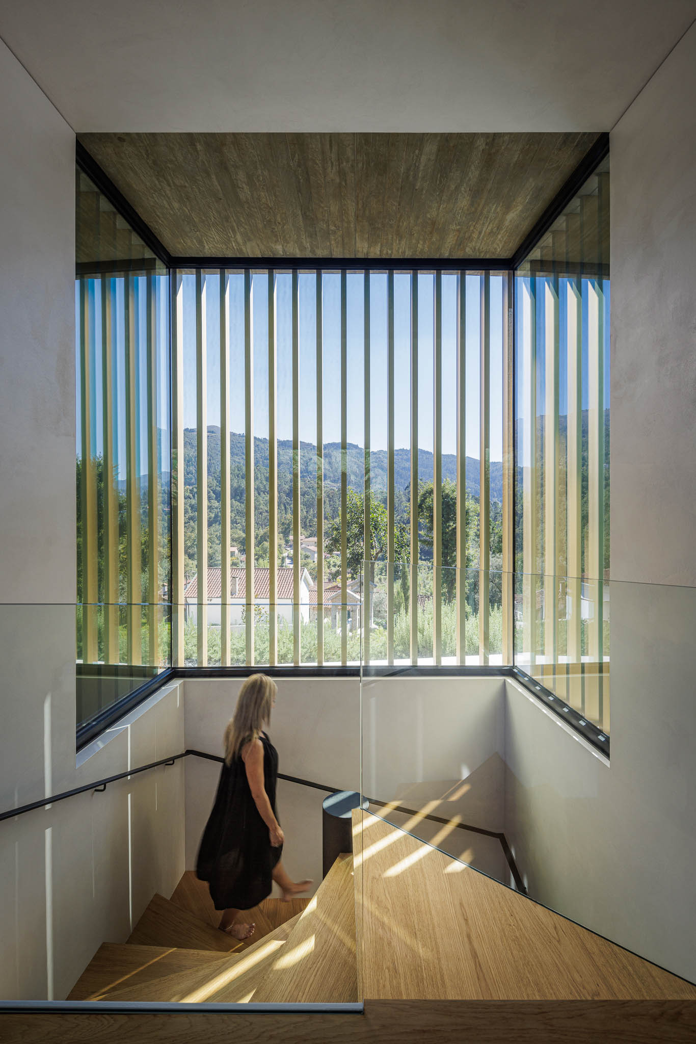 Escadas: Vista vertical da escada com ripas douradas ao fundo e mulher a descer os degraus de madeira. — Casa de Bouro arquitetura Mutant Architecture and Design em Terras de Bouro