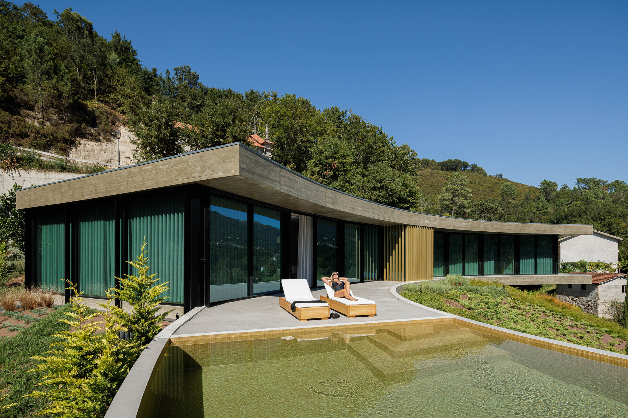 Exterior: Fachada curva da casa com piscina, espreguiçadeiras e jardim, integrando-se na paisagem natural. — Casa de Bouro arquitetura Mutant Architecture and Design em Terras de Bouro