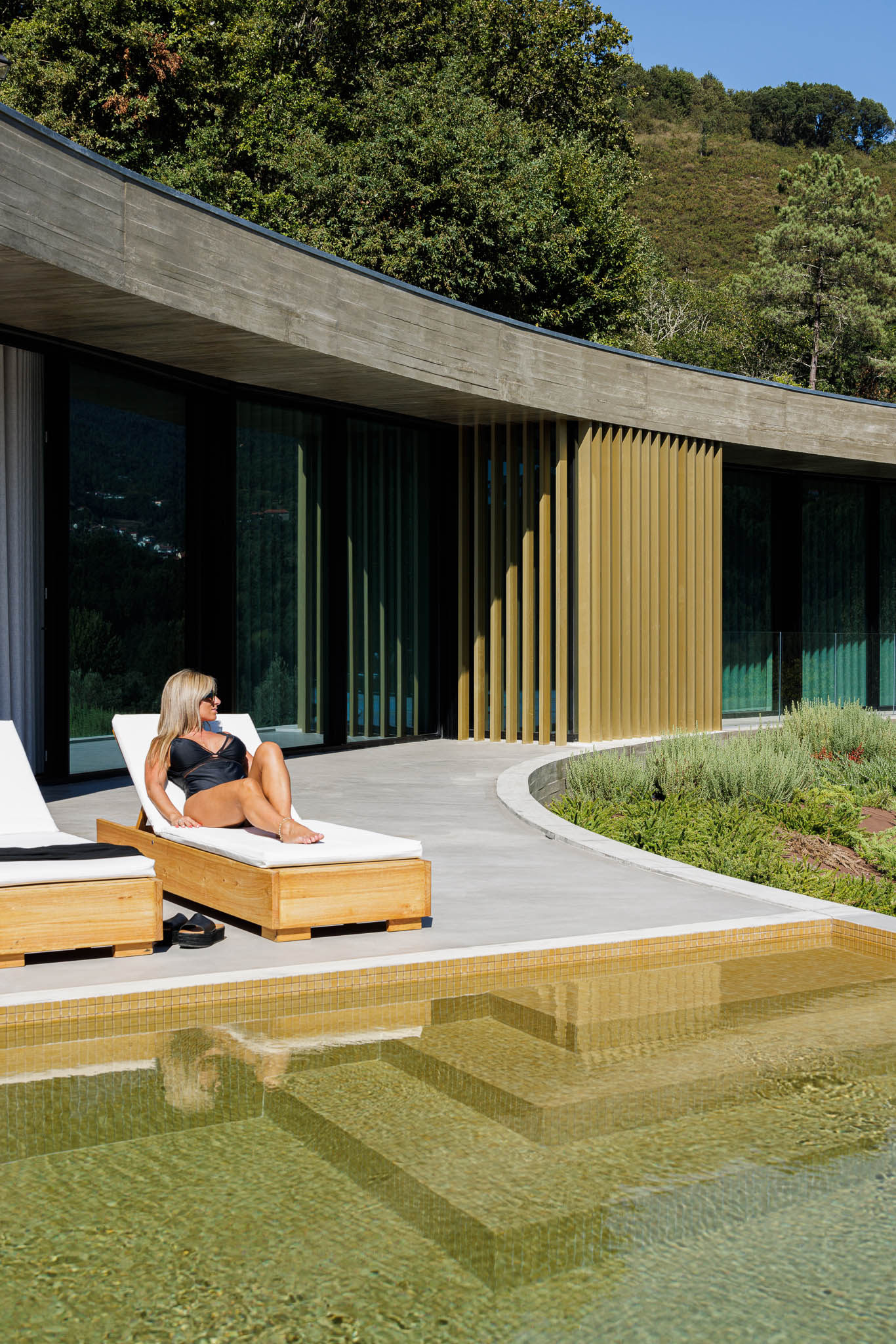 Exterior: Detalhe da piscina com degraus, espreguiçadeiras e fachada da casa com ripas douradas. — Casa de Bouro arquitetura Mutant Architecture and Design em Terras de Bouro
