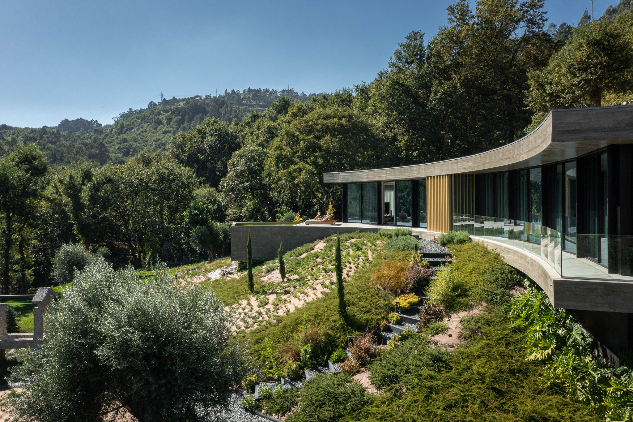 Exterior: Casa curva em betão e vidro, com jardim em socalcos e vegetação densa. — Casa de Bouro arquitetura Mutant Architecture and Design em Terras de Bouro