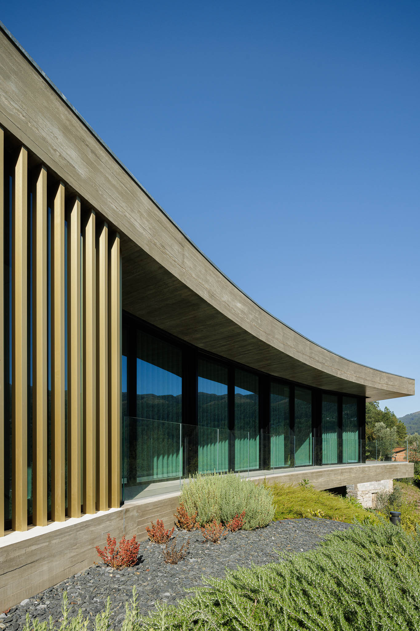 Exterior: Detalhe da fachada curva com ripas douradas, janelas amplas e jardim com vegetação rasteira. — Casa de Bouro arquitetura Mutant Architecture and Design em Terras de Bouro