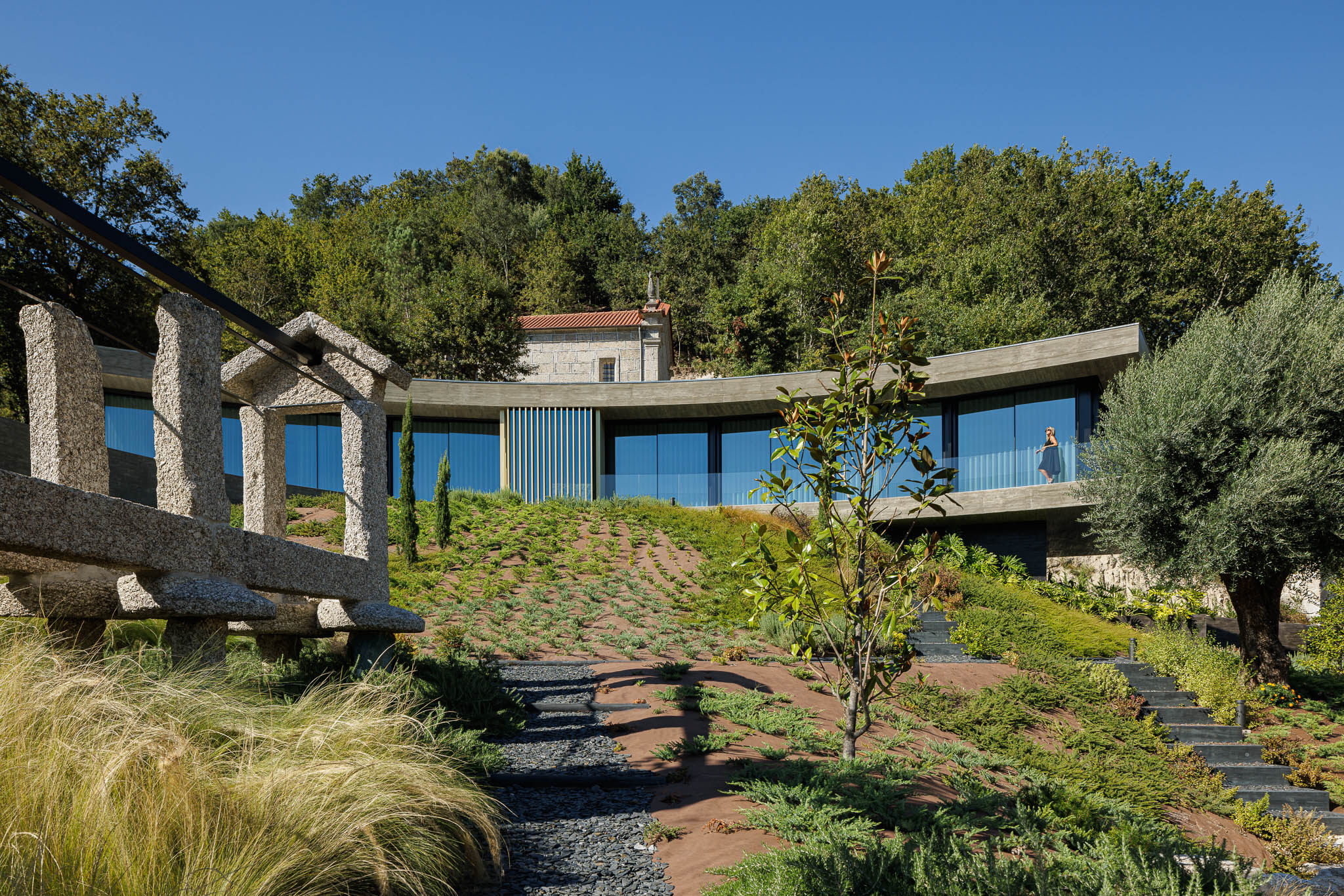 Exterior: Casa curva com fachada envidraçada, jardim em socalcos e estrutura antiga em pedra. — Casa de Bouro arquitetura Mutant Architecture and Design em Terras de Bouro