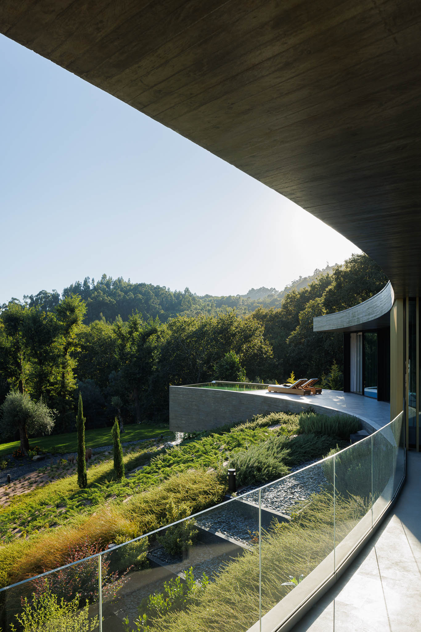 Exterior: Varanda curva com guarda-corpo em vidro, vista para a piscina e jardim em socalcos. — Casa de Bouro arquitetura Mutant Architecture and Design em Terras de Bouro