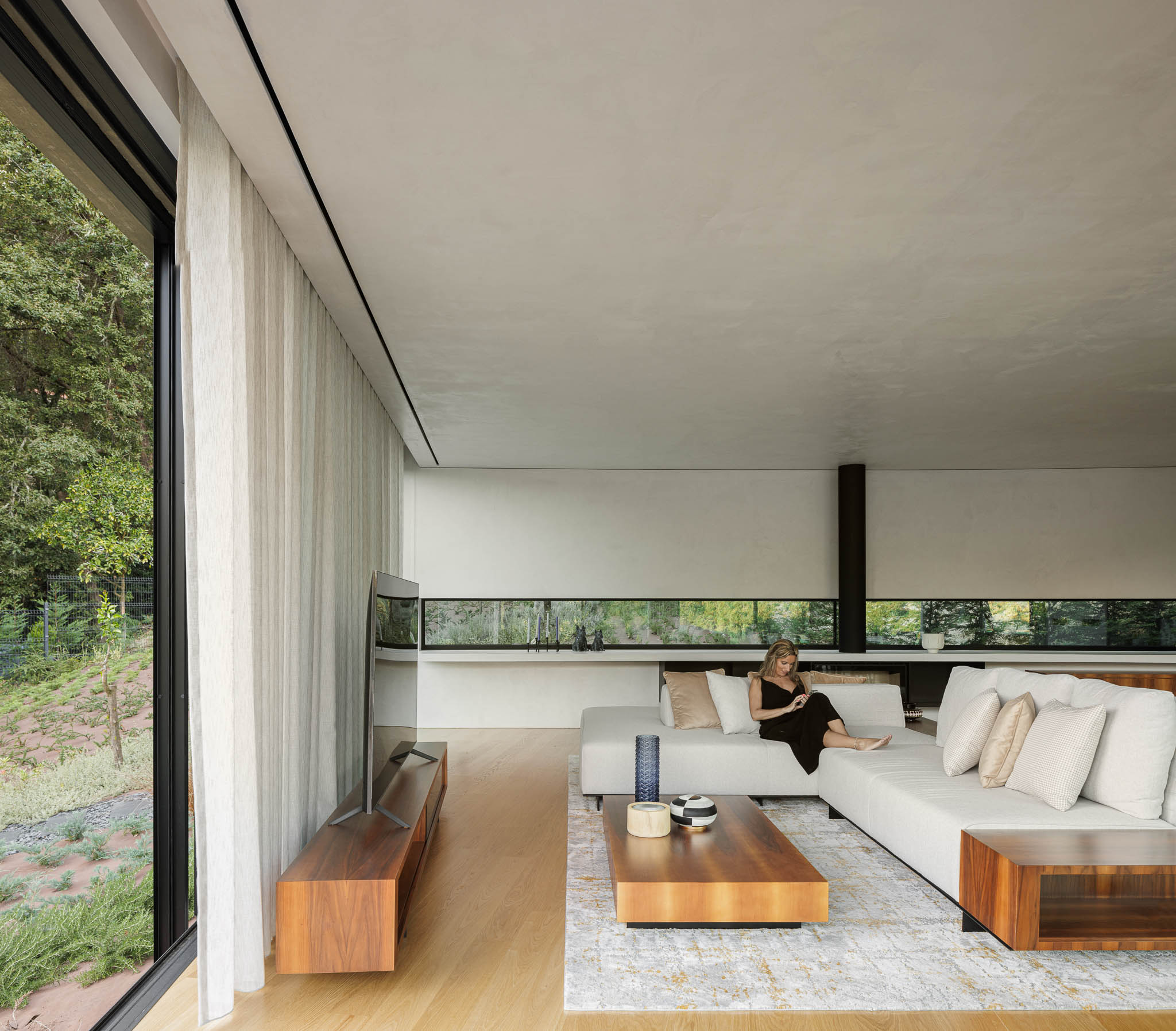 Sala de estar: Sala de estar com sofá branco, mesa de centro em madeira e grandes janelas com vista para o jardim. — Casa de Bouro arquitetura Mutant Architecture and Design em Terras de Bouro
