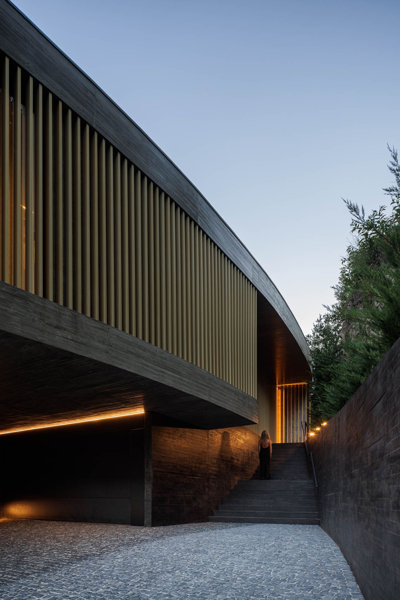 Exterior: Escadas de acesso à casa com fachada em betão e ripas douradas, iluminada por luz indireta. — Casa de Bouro arquitetura Mutant Architecture and Design em Terras de Bouro