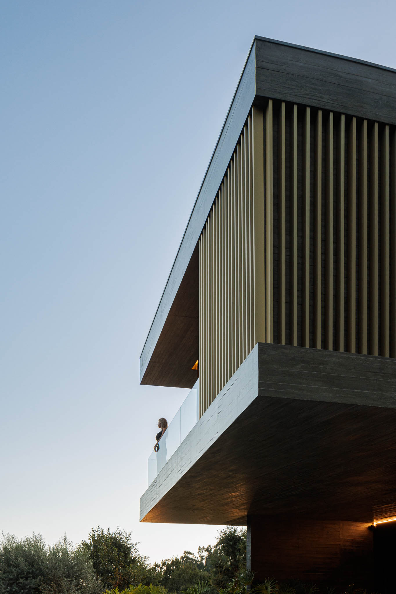 Exterior: Detalhe da fachada em betão com ripas douradas e varanda com guarda-corpo em vidro. — Casa de Bouro arquitetura Mutant Architecture and Design em Terras de Bouro