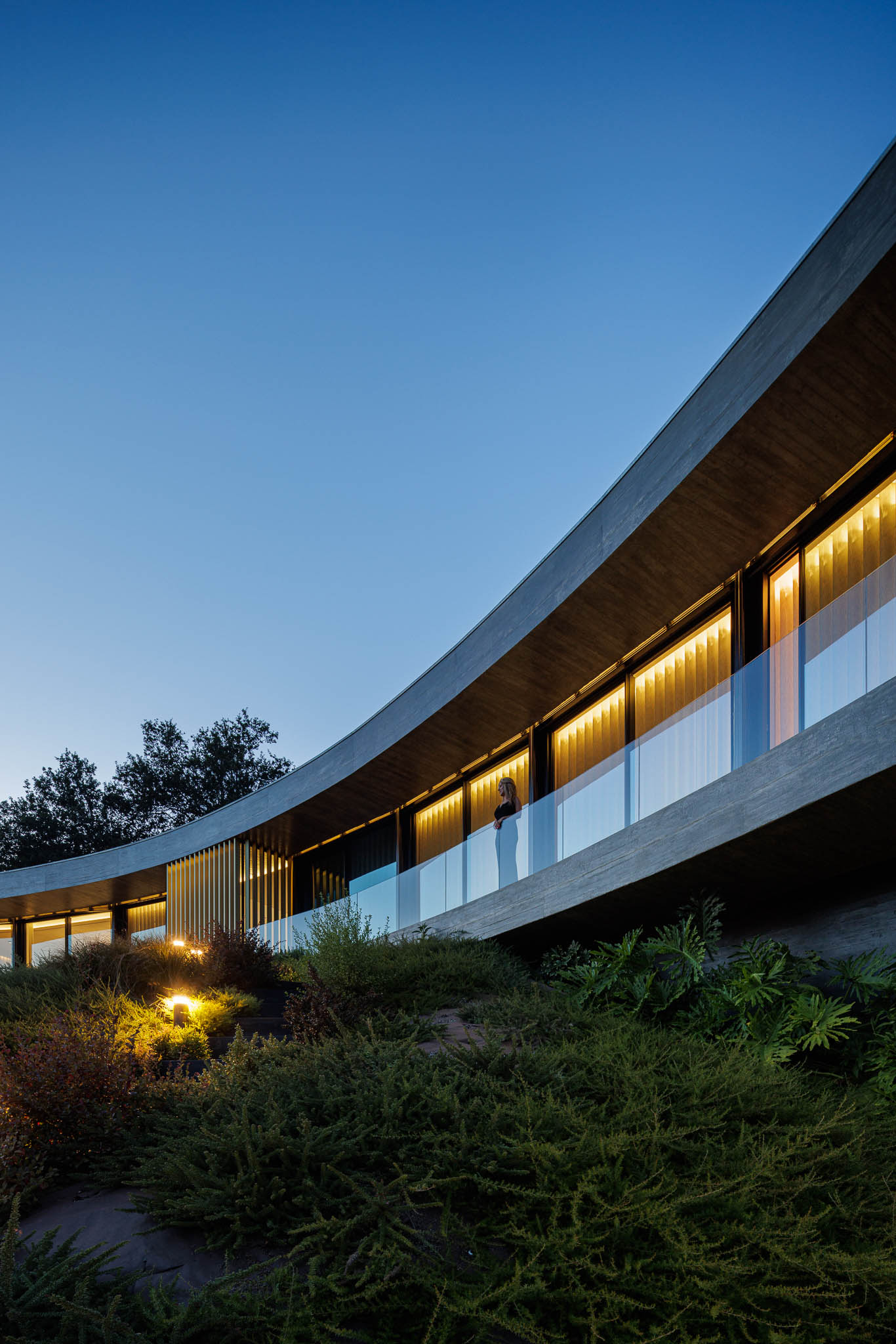 Exterior: Fachada curva da casa iluminada ao entardecer, com varanda em vidro e jardim. — Casa de Bouro arquitetura Mutant Architecture and Design em Terras de Bouro