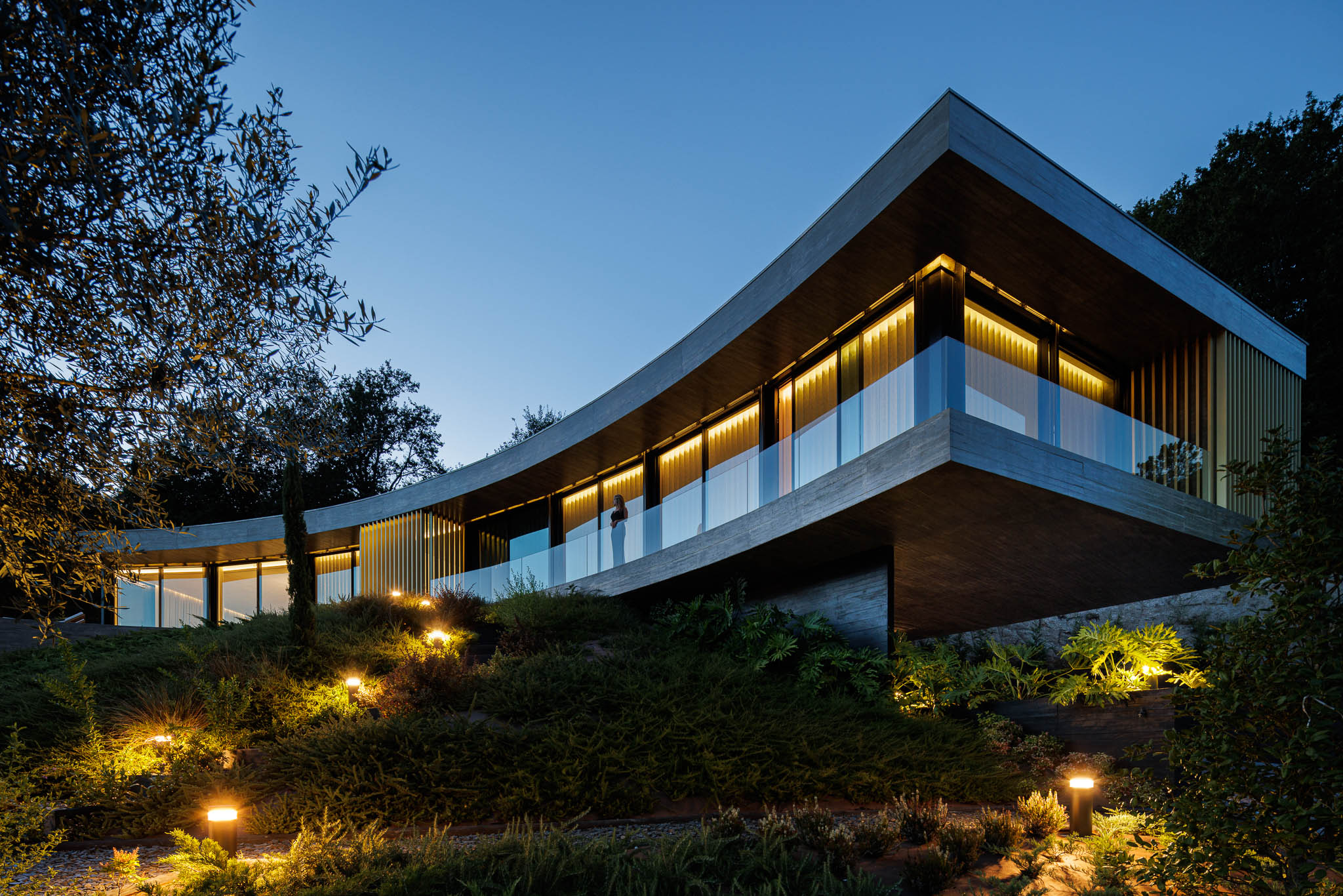Exterior: Vista noturna da casa com iluminação exterior, fachada curva e jardim em socalcos. — Casa de Bouro arquitetura Mutant Architecture and Design em Terras de Bouro