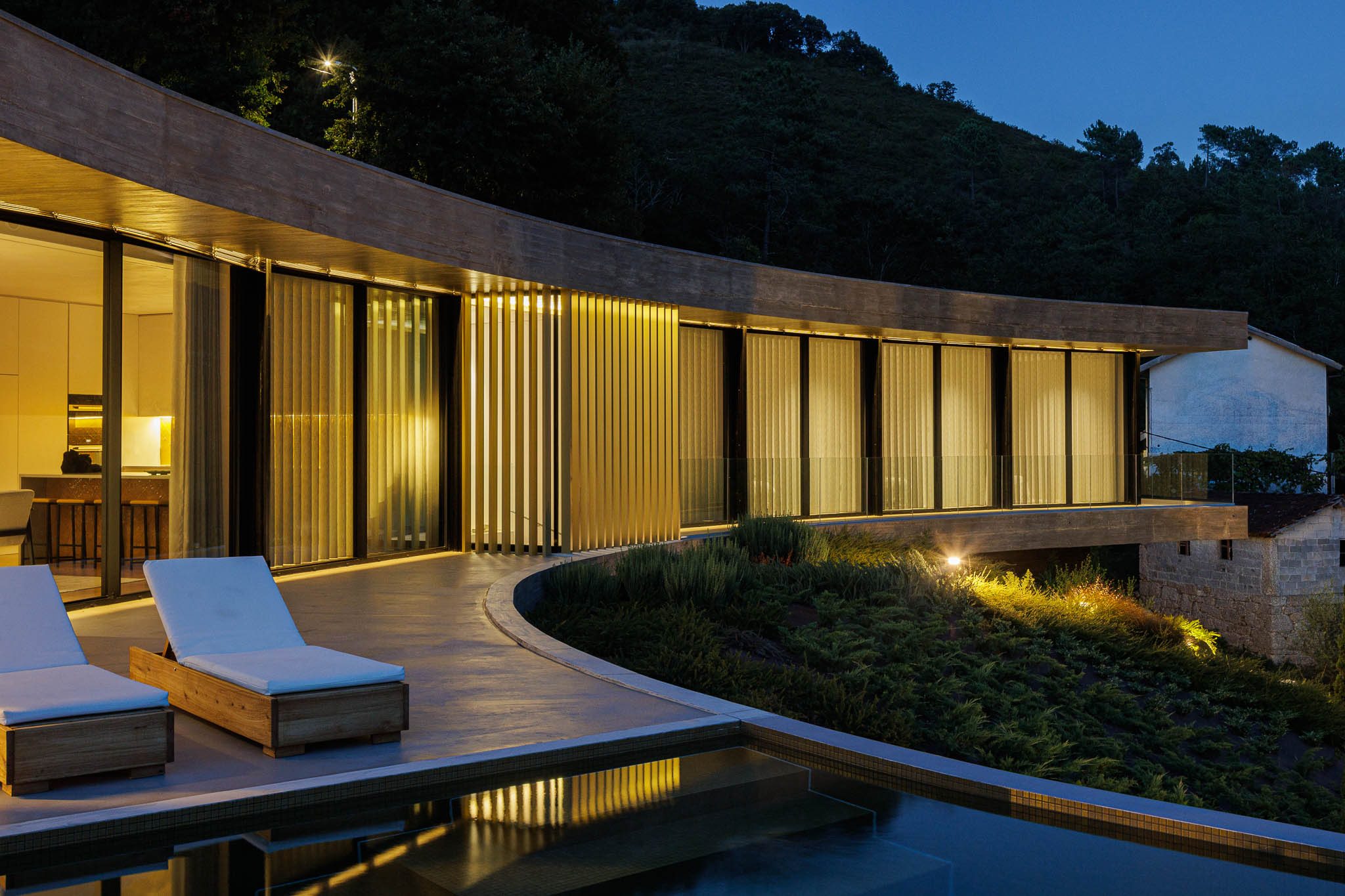 Exterior: Vista noturna da casa com piscina, espreguiçadeiras e fachada curva iluminada. — Casa de Bouro arquitetura Mutant Architecture and Design em Terras de Bouro