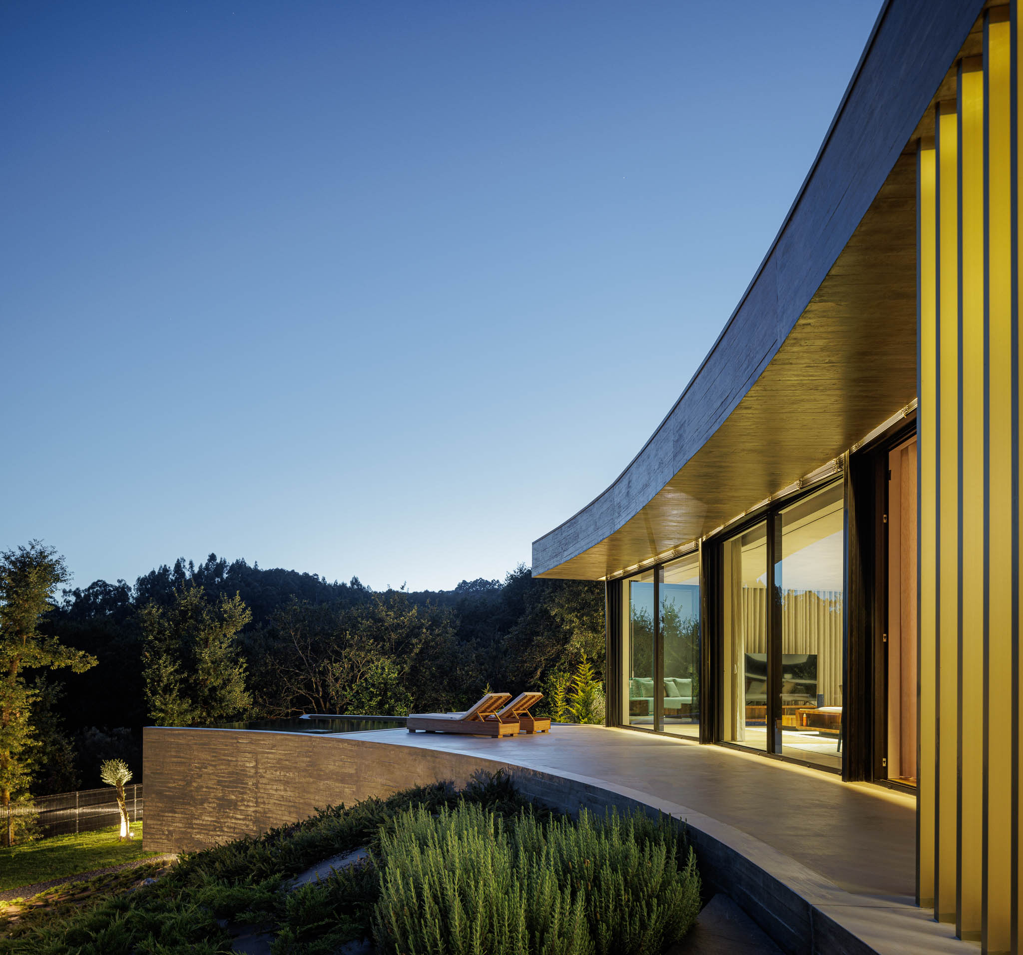 Exterior: Fachada curva da casa iluminada ao entardecer, com piscina e jardim. — Casa de Bouro arquitetura Mutant Architecture and Design em Terras de Bouro