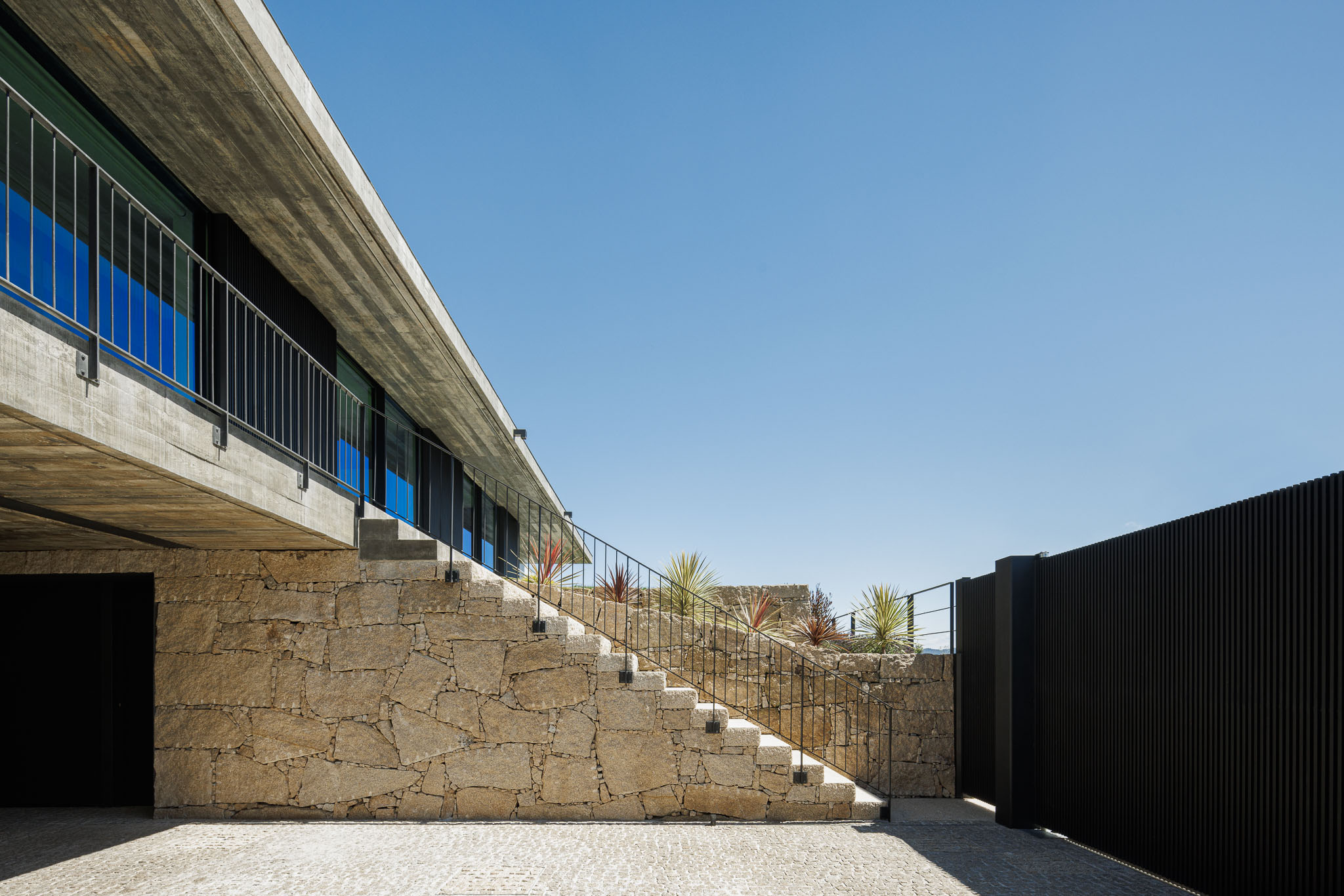 Acesso: Escadaria exterior em pedra rústica, com corrimão metálico preto, conduzindo à entrada principal da casa. Muro em pedra e portão metálico. — Casa Ponte arquitetura stu.dere em Amarante