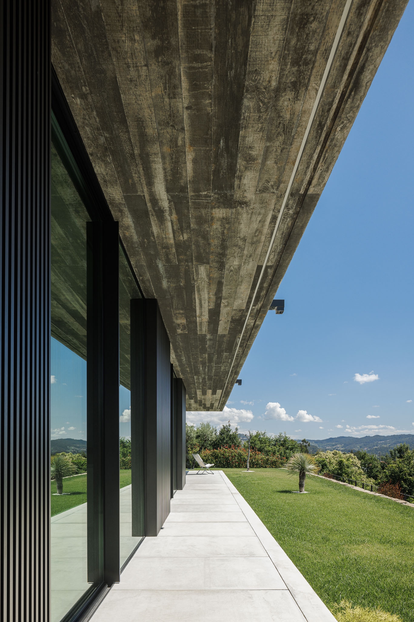 Alpendre: Teto em betão aparente, pilares metálicos pretos e pavimento em betão liso. Vista para a paisagem verdejante. — Casa Ponte arquitetura stu.dere em Amarante