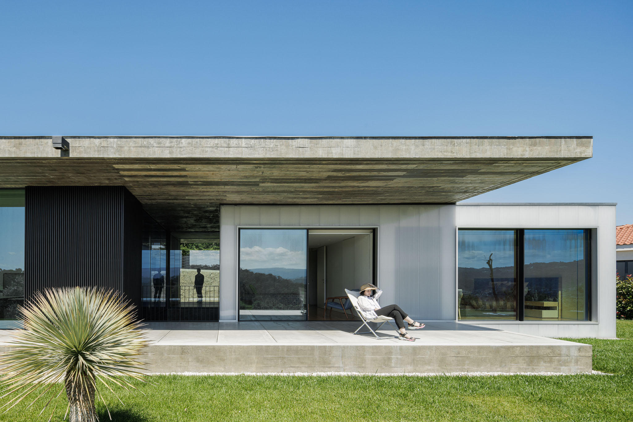 Alpendre: Fachada com revestimento translúcido e estrutura em betão. Pessoa sentada numa cadeira de exterior. — Casa Ponte arquitetura stu.dere em Amarante