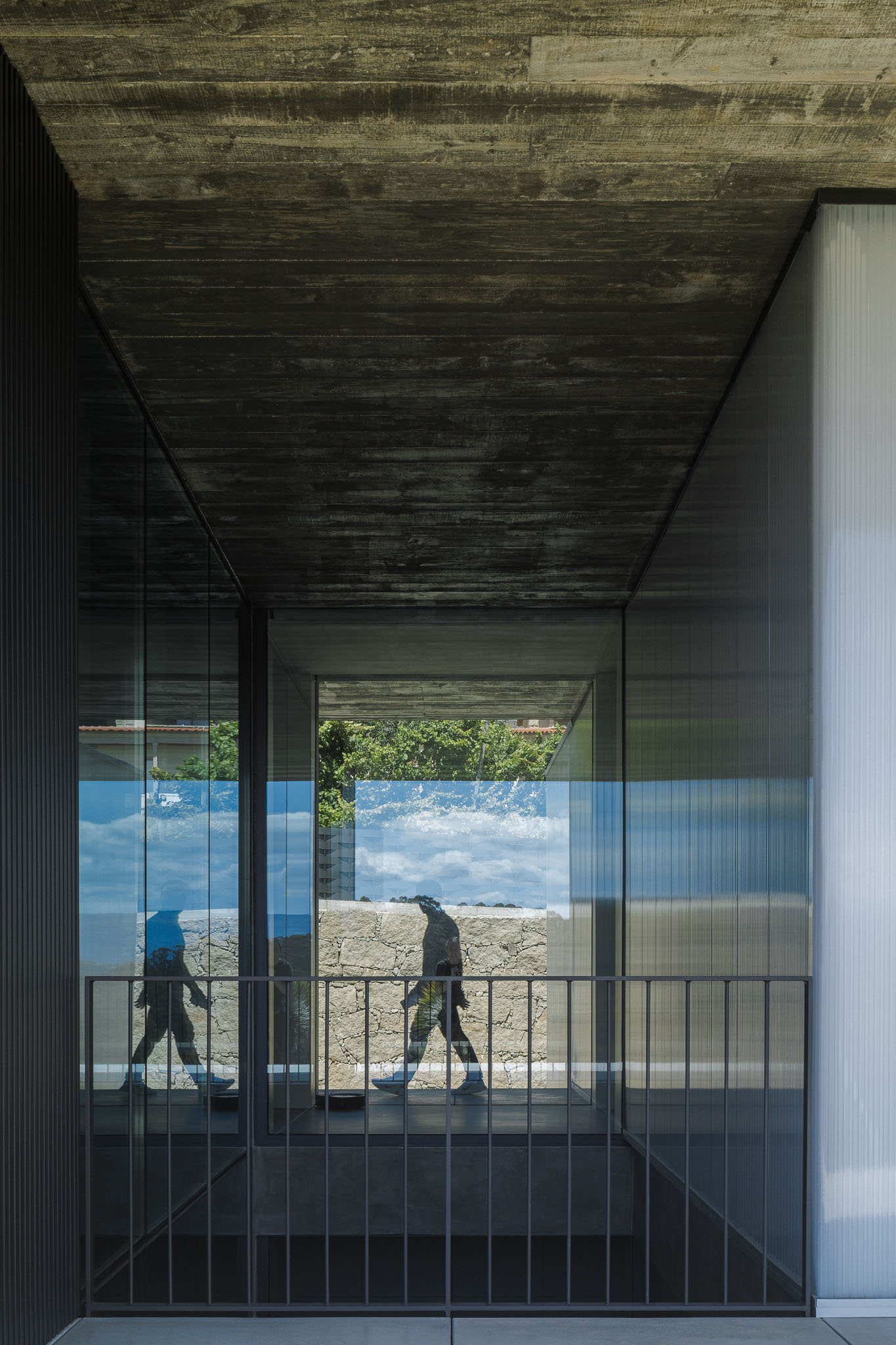 Circulação: Corredor envidraçado com teto em betão aparente e pavimento em betão. Reflexo da paisagem no vidro. — Casa Ponte arquitetura stu.dere em Amarante