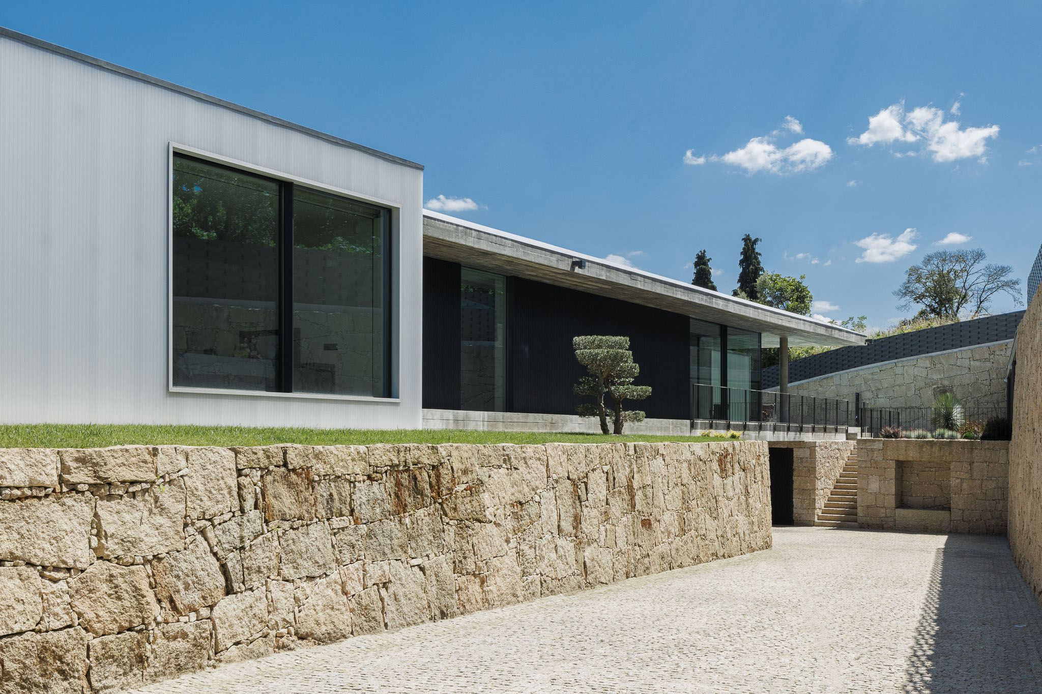 Acesso: Muro em pedra rústica e casa com revestimento translúcido e madeira escura. Escadaria em pedra. — Casa Ponte arquitetura stu.dere em Amarante