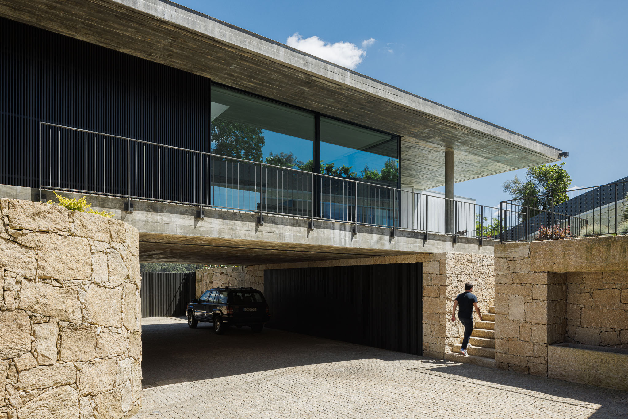 Garagem: Entrada para a garagem sob a casa, com portão metálico preto. Escadaria em pedra e muro rústico. — Casa Ponte arquitetura stu.dere em Amarante