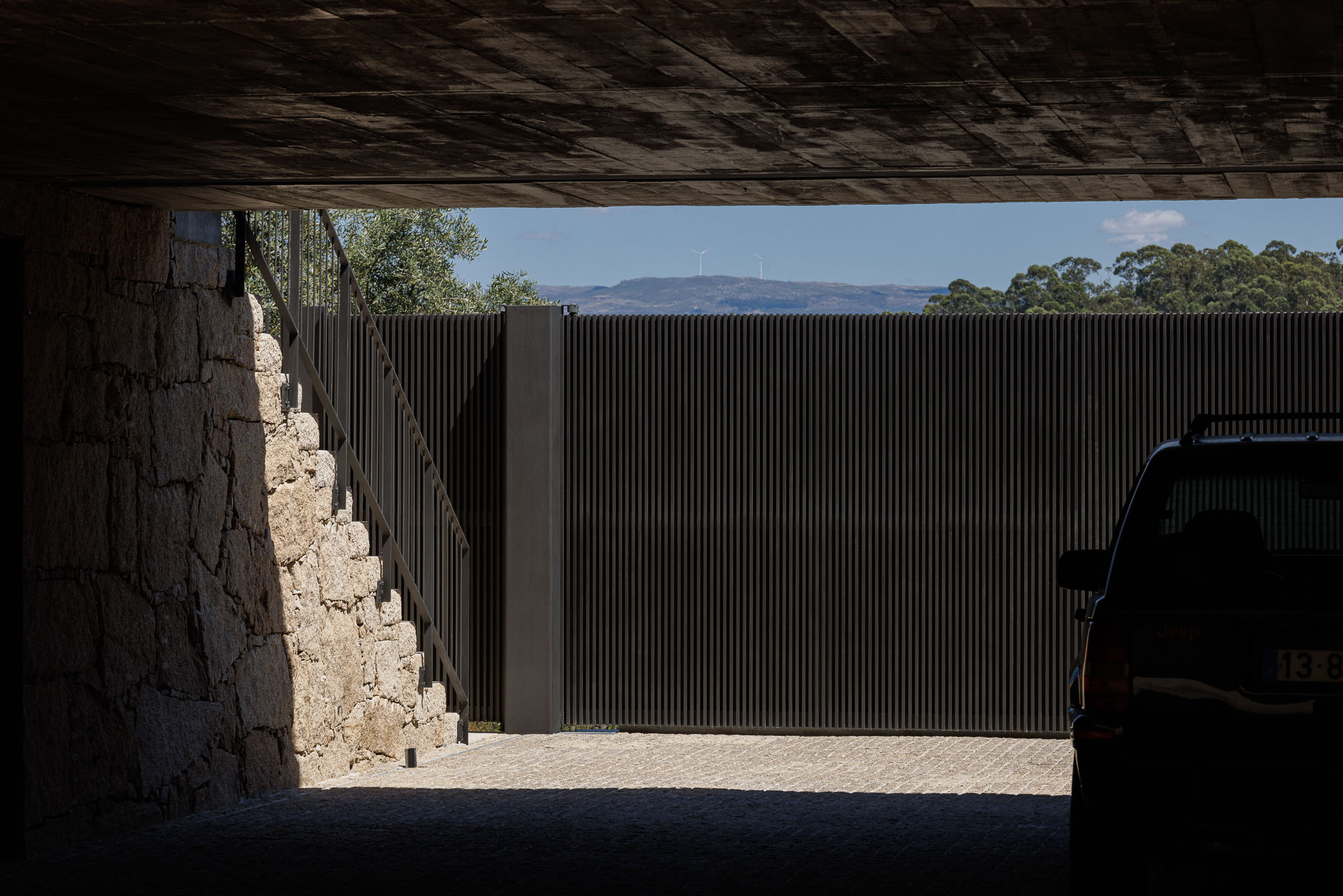 Garagem: Interior da garagem com portão metálico preto e pavimento em calçada. Muro em pedra rústica. — Casa Ponte arquitetura stu.dere em Amarante