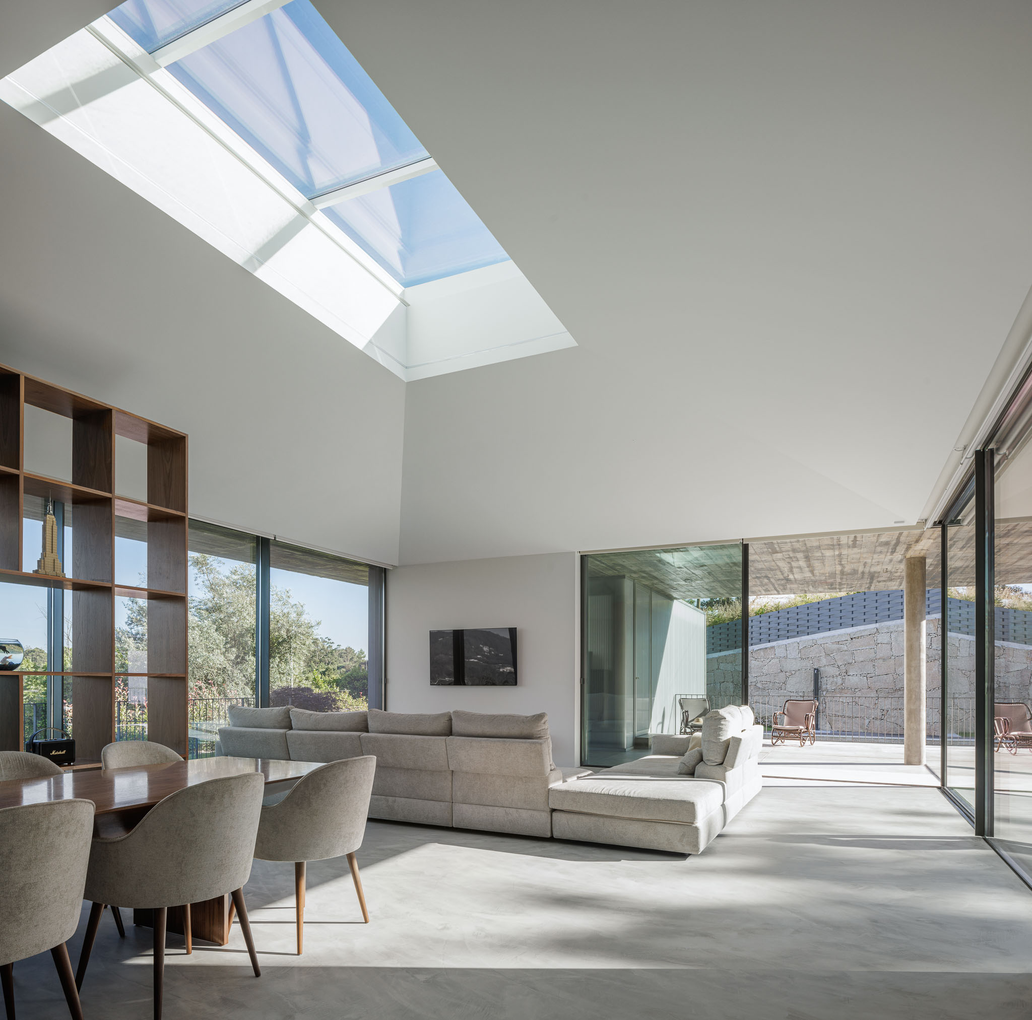 Sala de estar: Espaço amplo com sofá modular, estante em madeira e janelas panorâmicas. Teto alto com claraboia. — Casa Ponte arquitetura stu.dere em Amarante