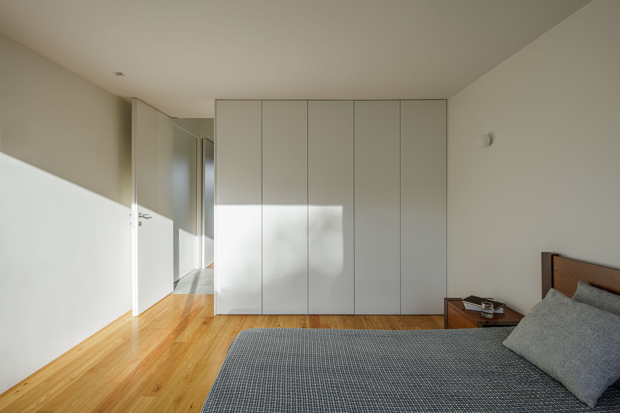 Quarto: Cama com roupa de cama cinzenta, armário embutido branco e pavimento em madeira. — Casa Ponte arquitetura stu.dere em Amarante