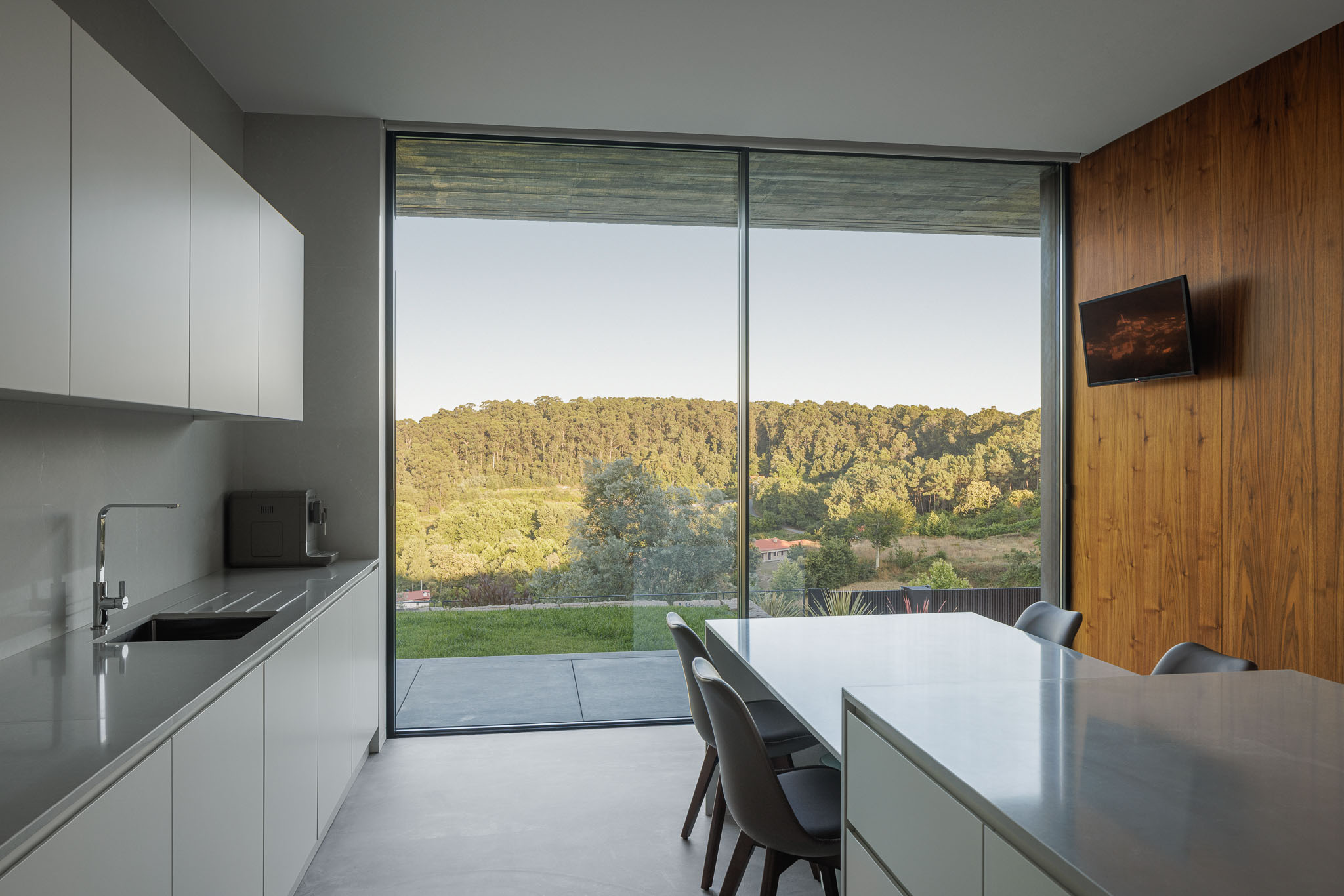 Cozinha: Armários brancos, bancada em pedra e revestimento em madeira. Janela panorâmica com vista para o exterior. — Casa Ponte arquitetura stu.dere em Amarante