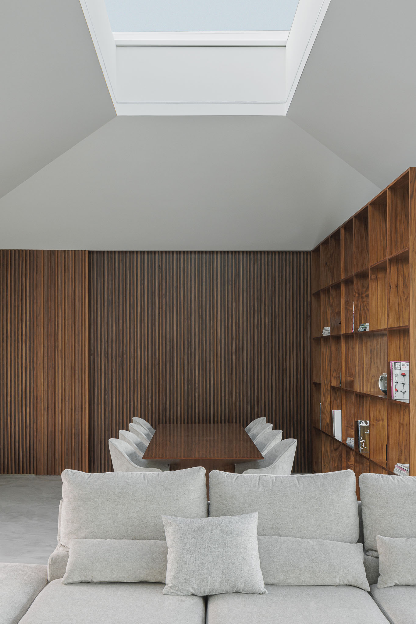 Sala de jantar: Mesa de jantar com cadeiras cinzentas, estante em madeira e divisória em madeira. Sofá em segundo plano. — Casa Ponte arquitetura stu.dere em Amarante