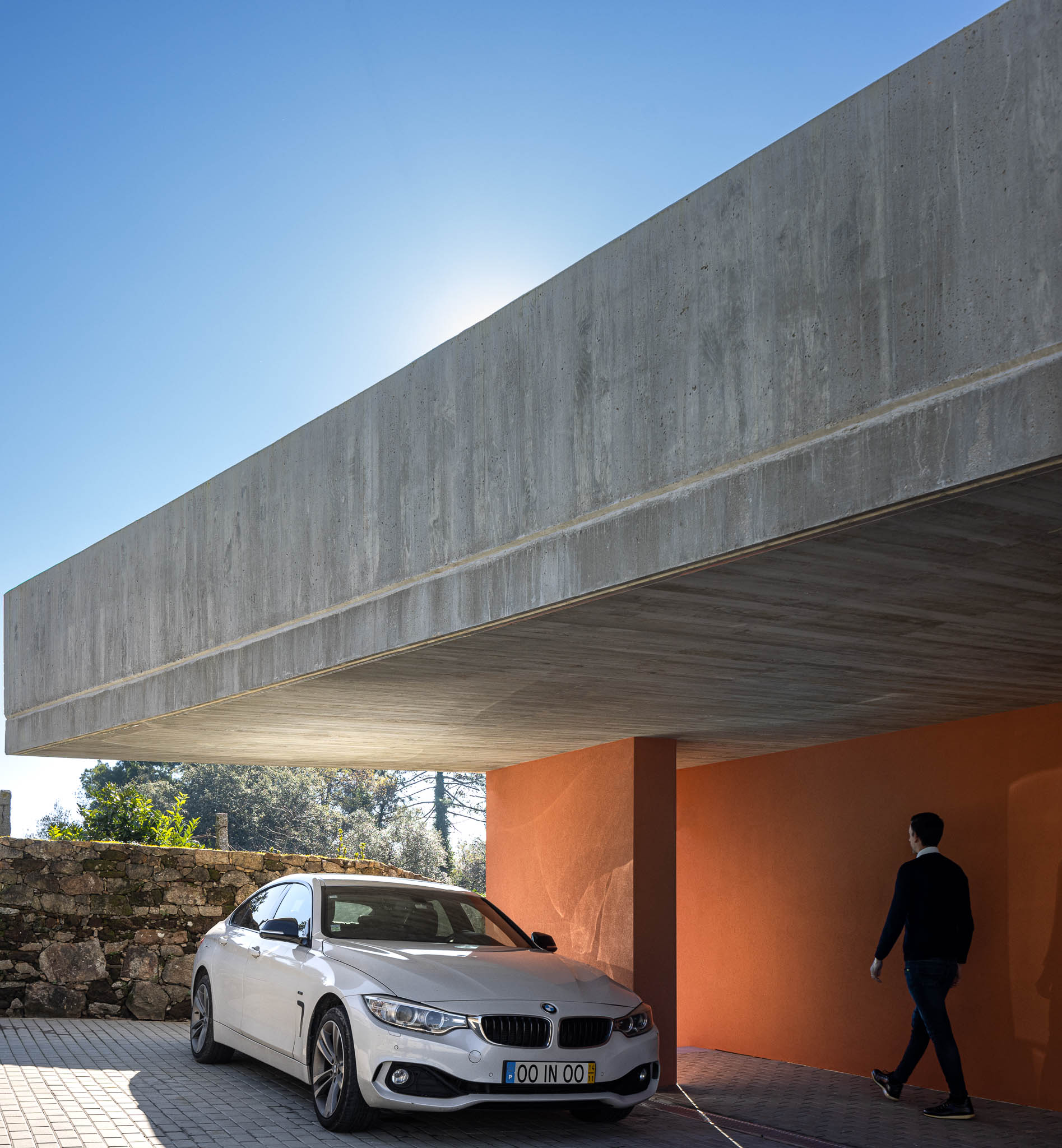 Alpendre: Laje de betão aparente, pilar cor de tijolo, pavimento em calçada. Carro estacionado. — TM House arquitetura Inception Architects Studio em Santo Tirso