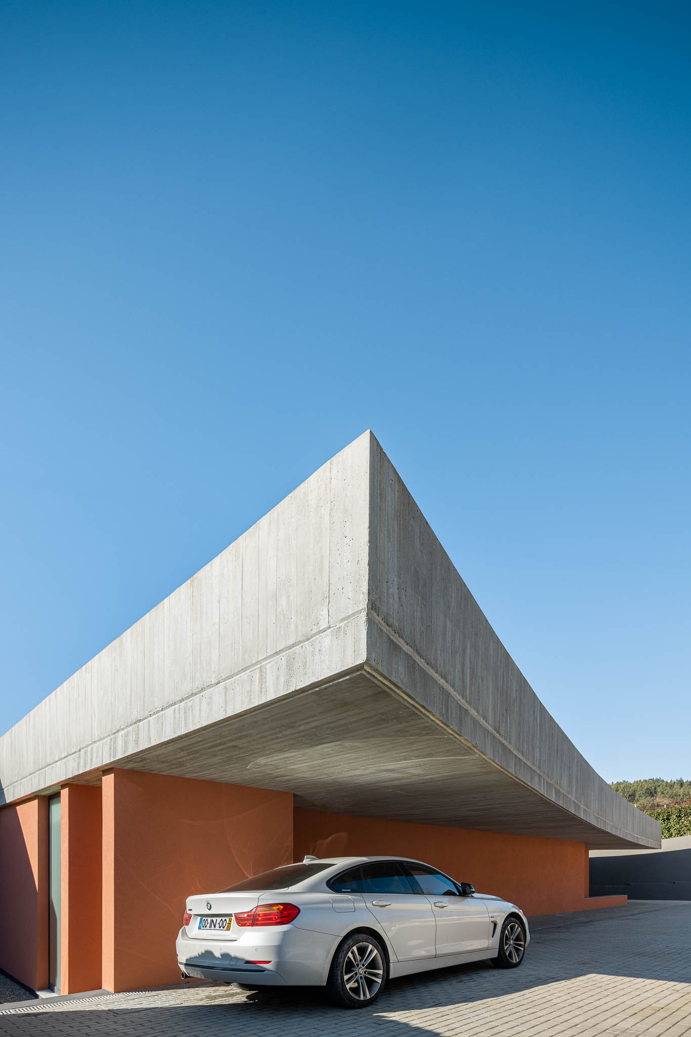 Exterior: Detalhe da esquina da casa, laje de betão aparente e paredes cor de tijolo. Carro estacionado. — TM House arquitetura Inception Architects Studio em Santo Tirso