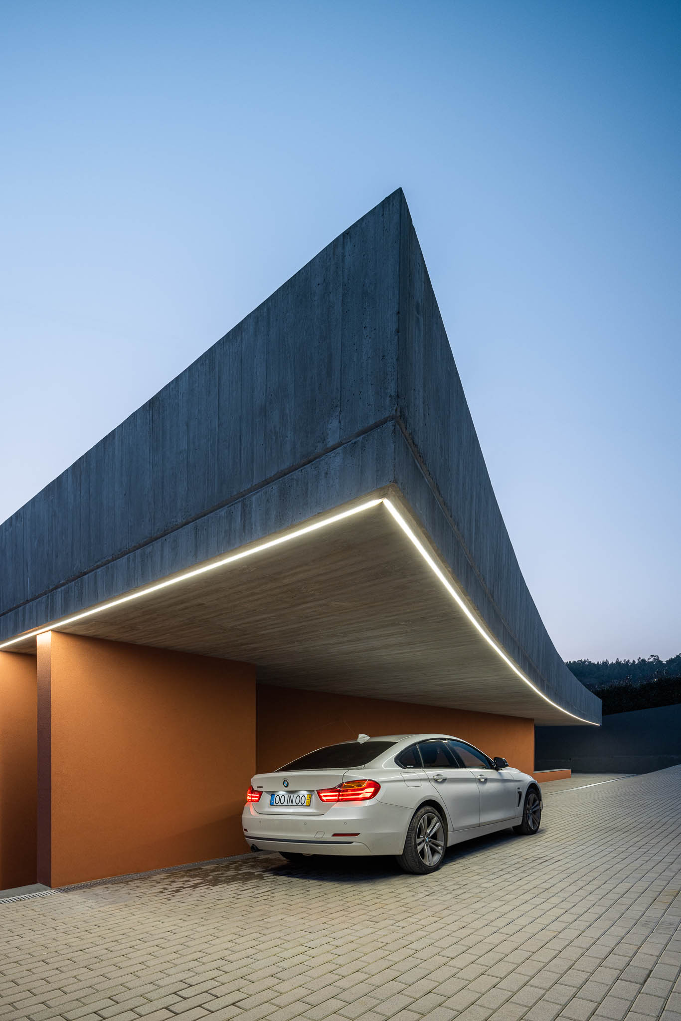 Exterior: Vista noturna da casa com laje de betão aparente com forma curva e paredes cor de tijolo. — TM House arquitetura Inception Architects Studio em Santo Tirso