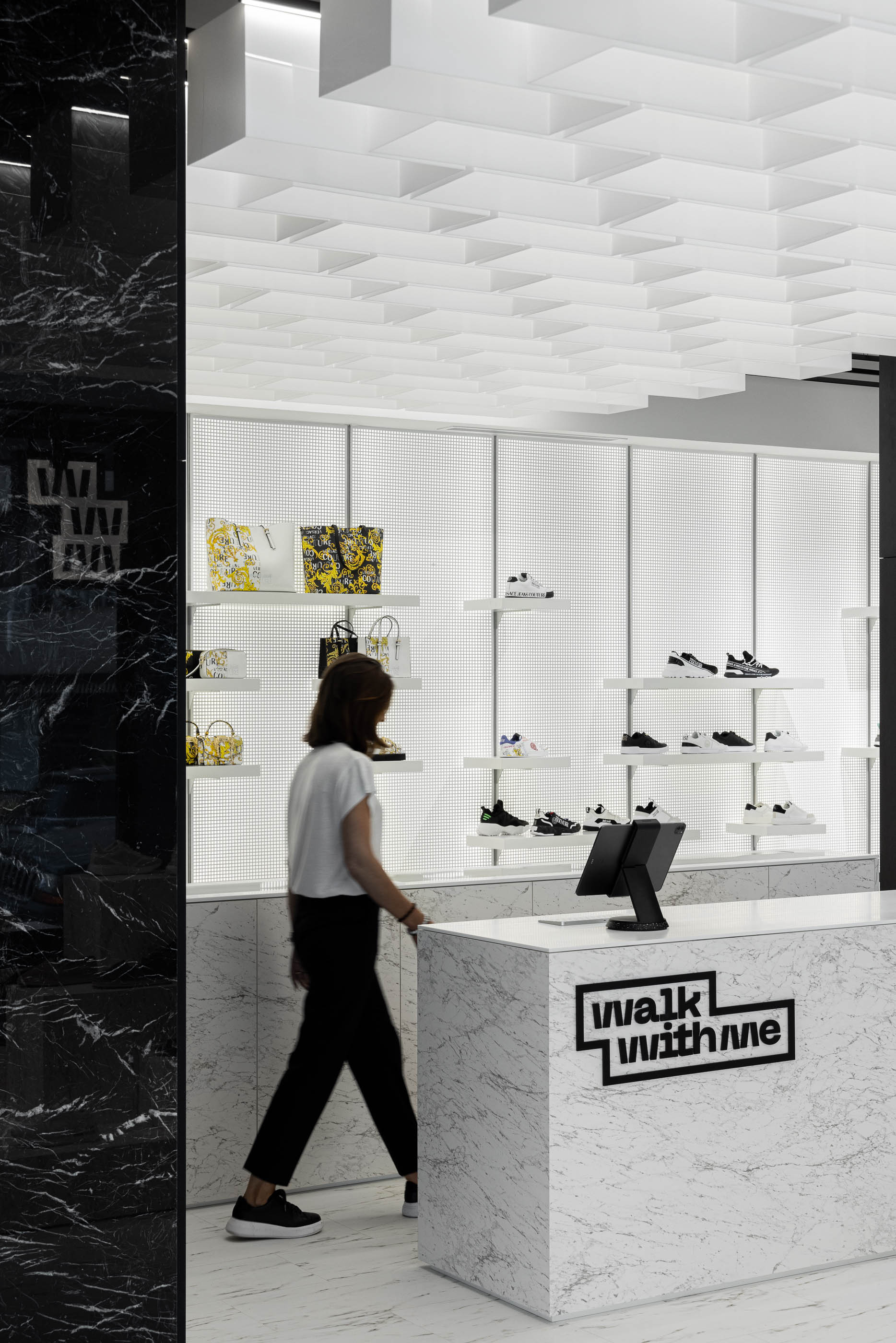 Loja: Balcão de atendimento em mármore branco com o logótipo, teto com elementos geométricos brancos e prateleiras. — Walk With Me Store arquitetura REM'A Arquitectos em Porto