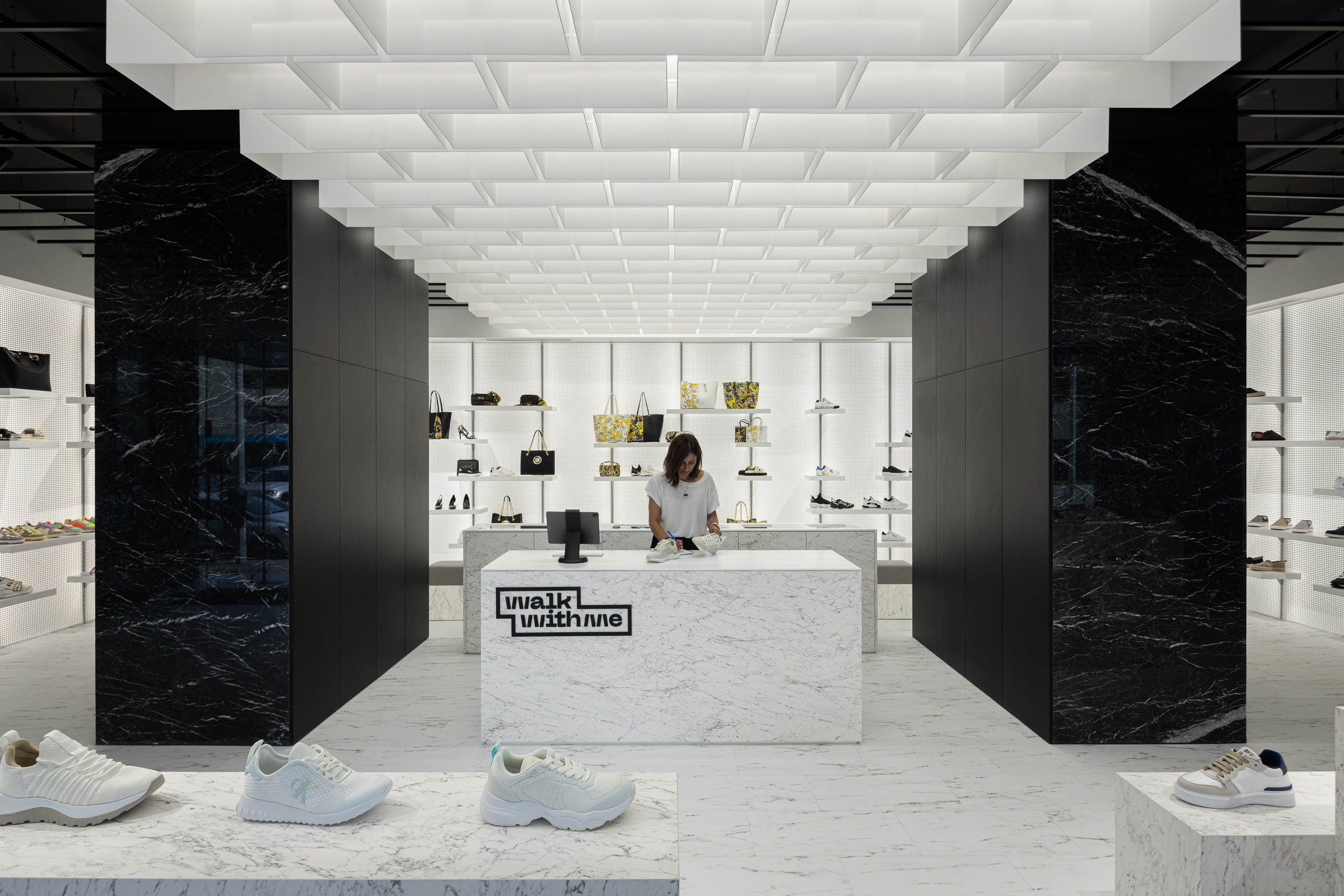 Loja: Perspectiva frontal do balcão em mármore, com teto geométrico e prateleiras laterais iluminadas. — Walk With Me Store arquitetura REM'A Arquitectos em Porto
