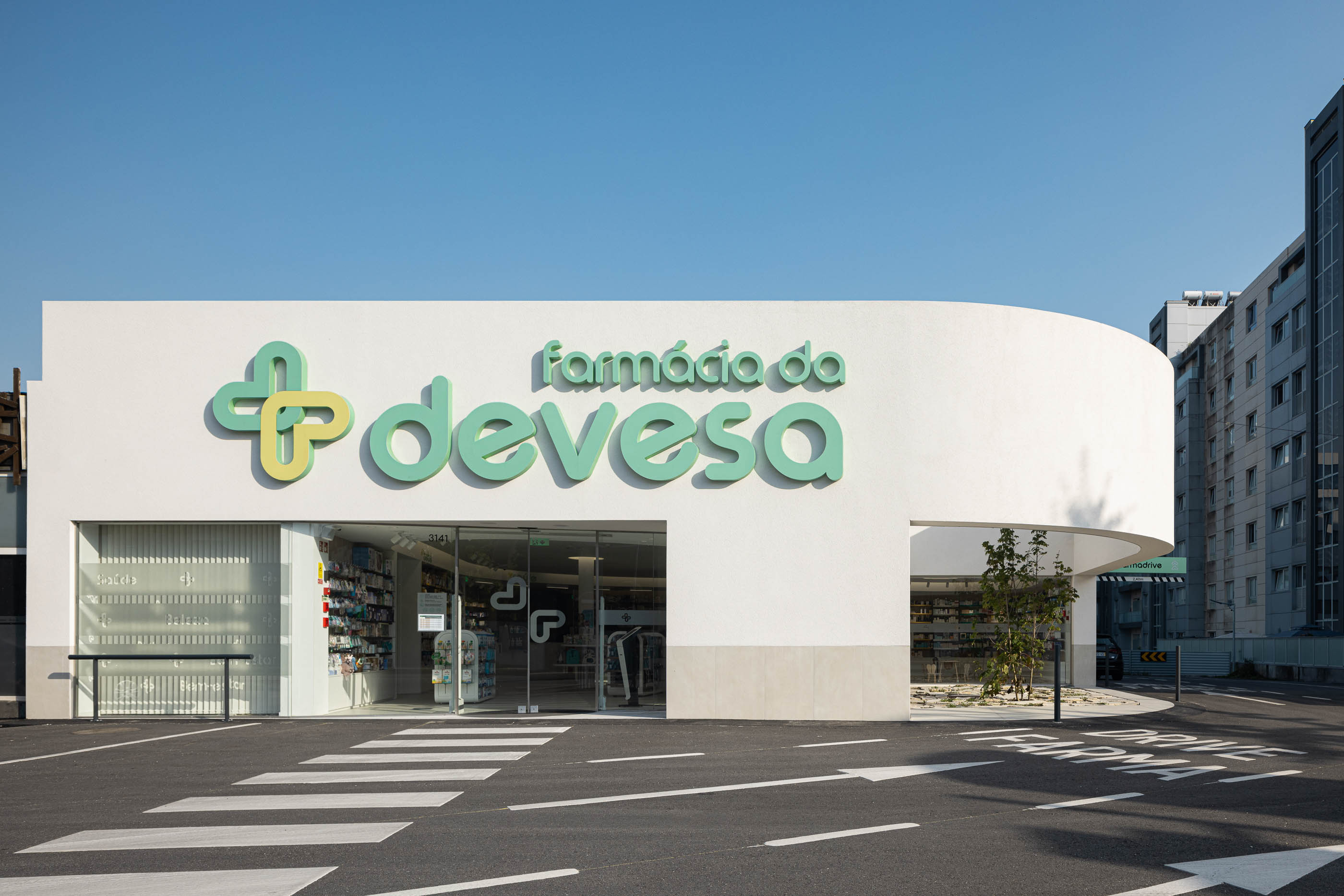 Entrada principal: Fachada branca curva com sinalização verde, estacionamento e passadeira. Arquitetura minimalista. — Farmácia da Devesa arquitetura Tsou Arquitectos em Vila Nova de Famalicão