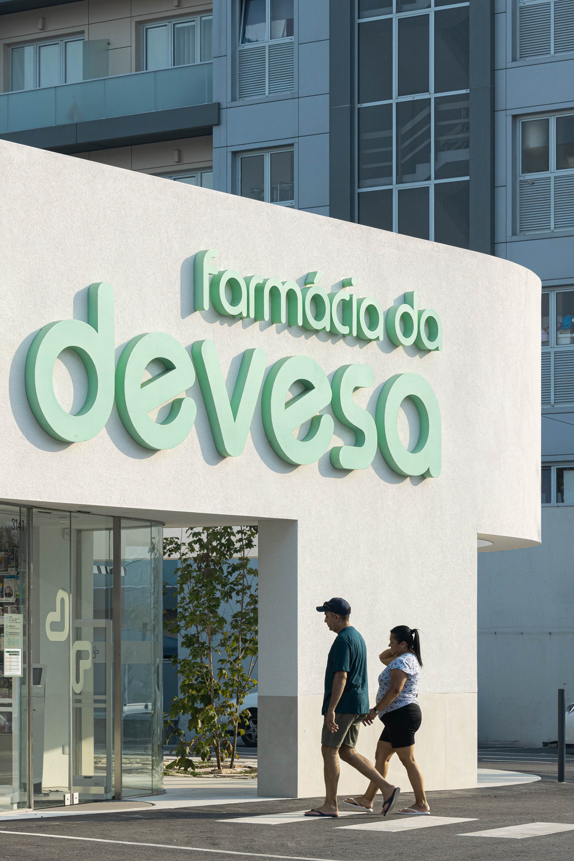 Entrada principal: Detalhe da fachada com a sinalização "Farmácia da Devesa" e pessoas a atravessar a passadeira. — Farmácia da Devesa arquitetura Tsou Arquitectos em Vila Nova de Famalicão