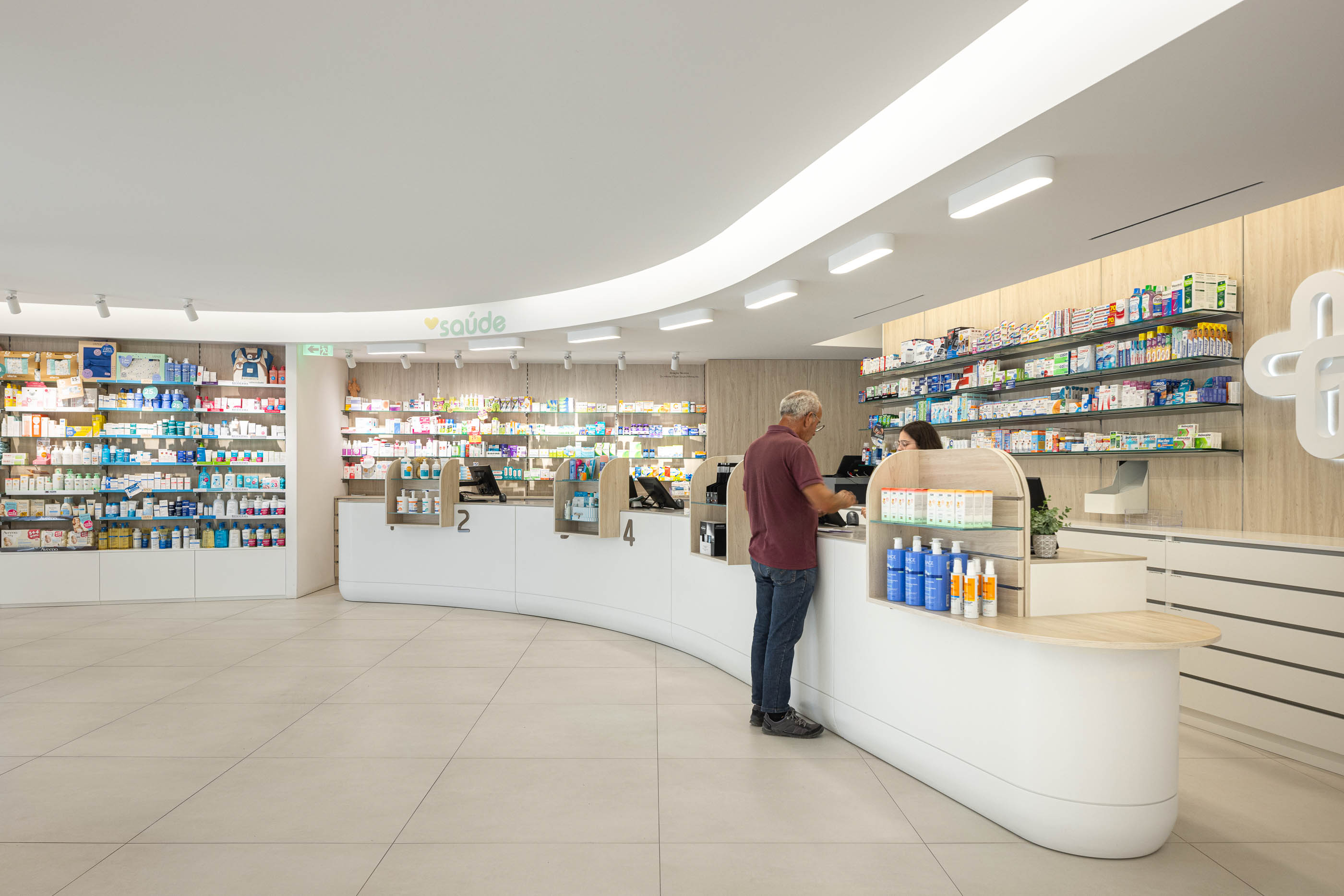 Área de atendimento: Balcão curvo branco com iluminação embutida e prateleiras com produtos. Pavimento cerâmico claro. — Farmácia da Devesa arquitetura Tsou Arquitectos em Vila Nova de Famalicão