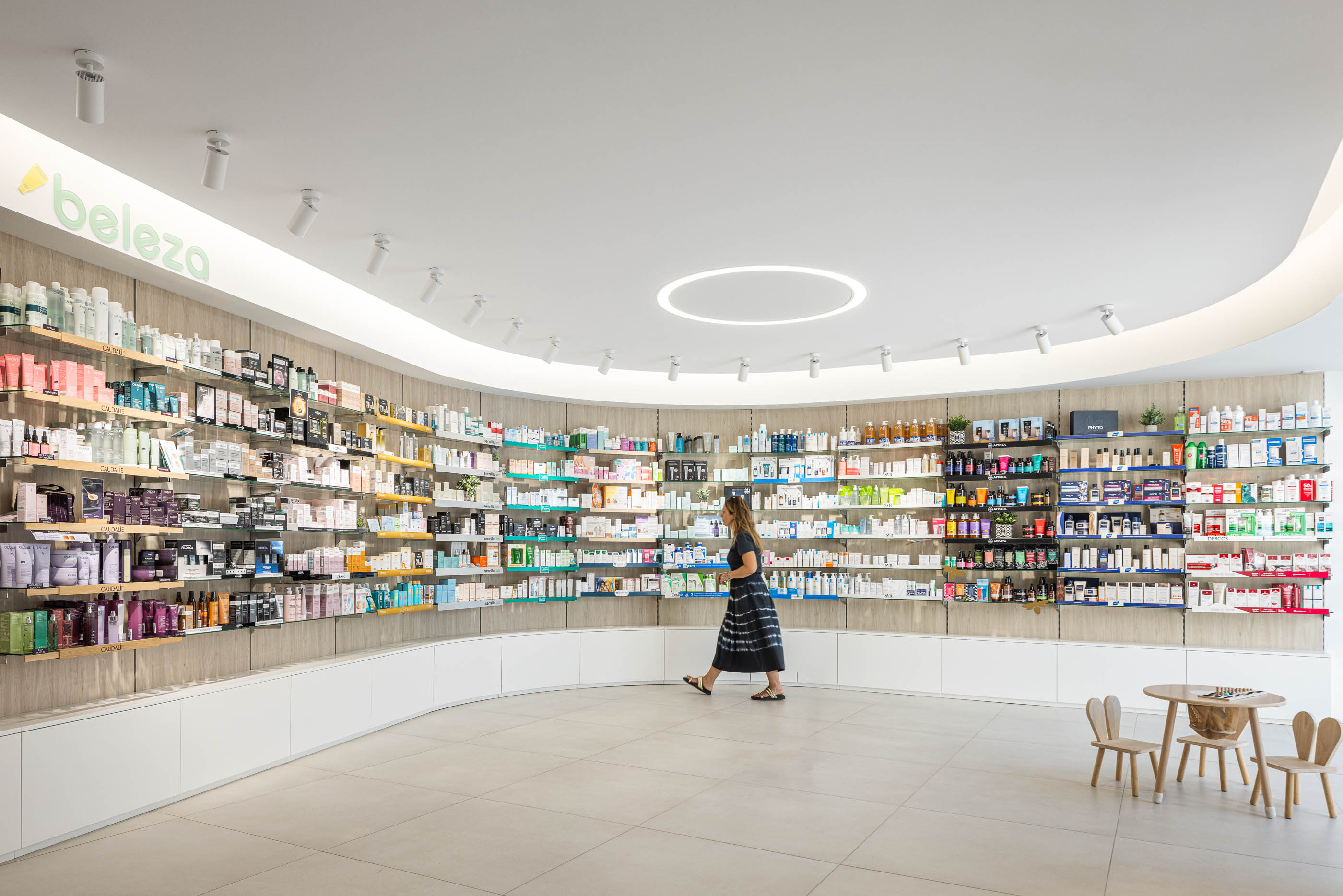 Área de beleza: Prateleiras curvas com produtos de beleza, pavimento cerâmico claro e candeeiros embutidos. — Farmácia da Devesa arquitetura Tsou Arquitectos em Vila Nova de Famalicão