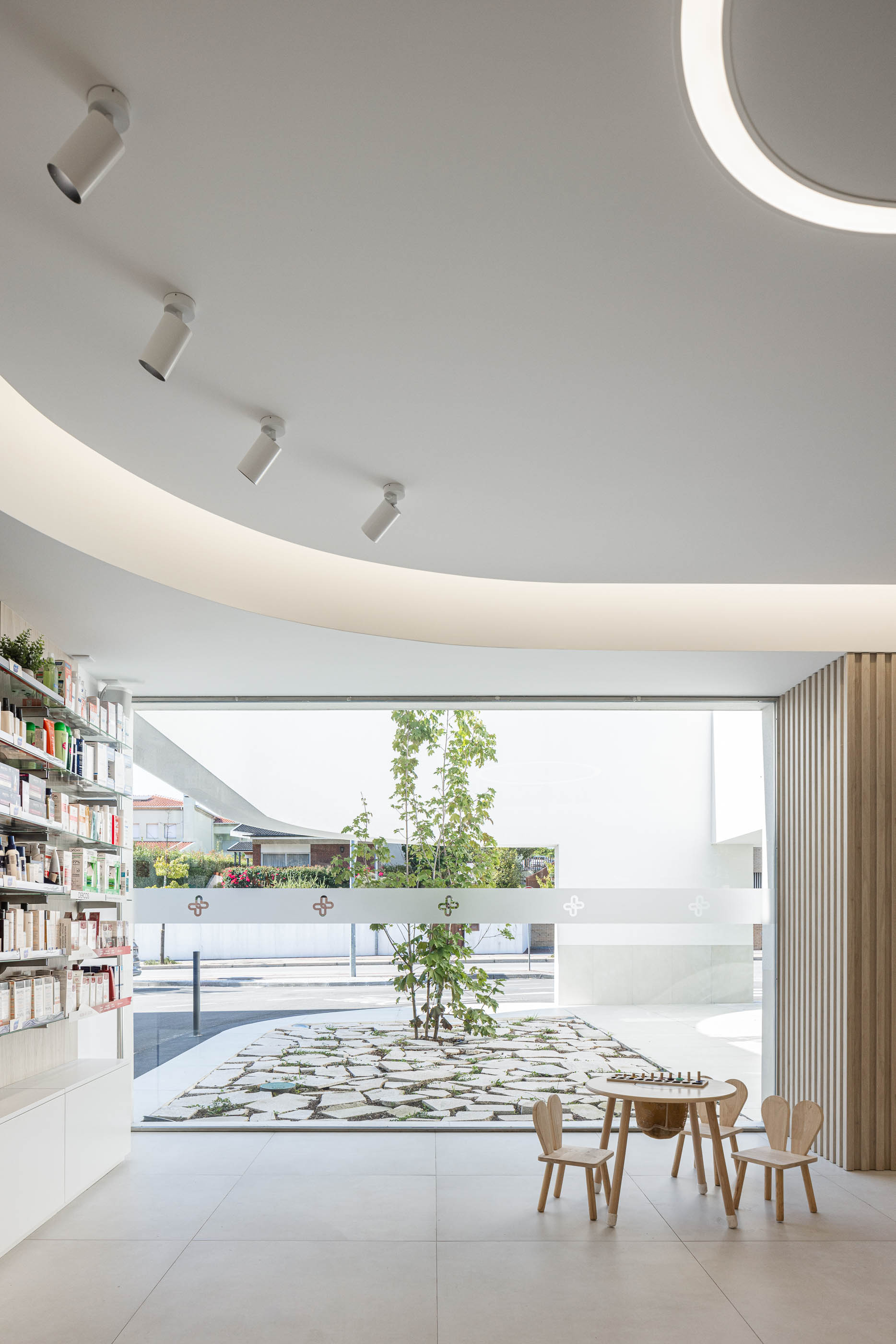 Área de espera: Mesa de jogos para crianças, com vista para o jardim interior e prateleiras com produtos. — Farmácia da Devesa arquitetura Tsou Arquitectos em Vila Nova de Famalicão