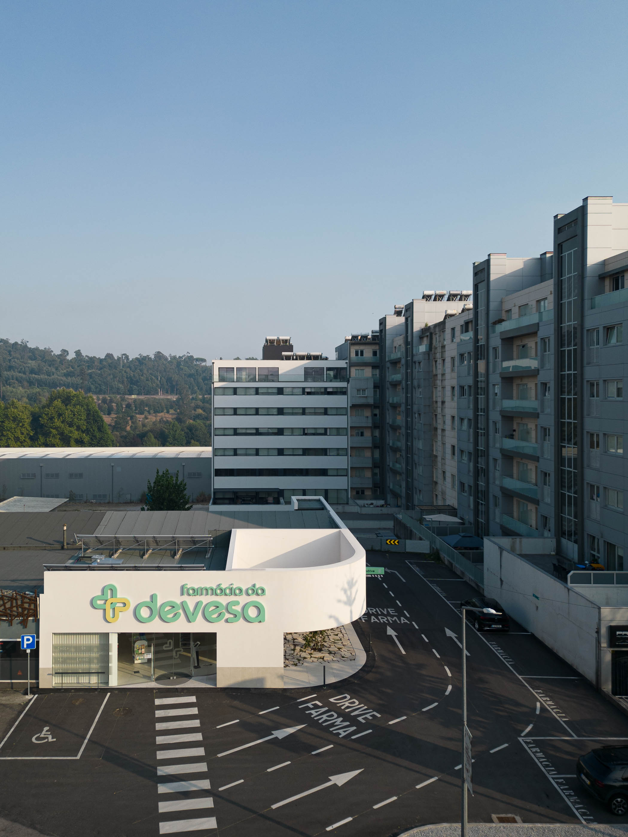 Exterior: Vista geral da farmácia, inserida no contexto urbano, com estacionamento e edifícios residenciais. — Farmácia da Devesa arquitetura Tsou Arquitectos em Vila Nova de Famalicão