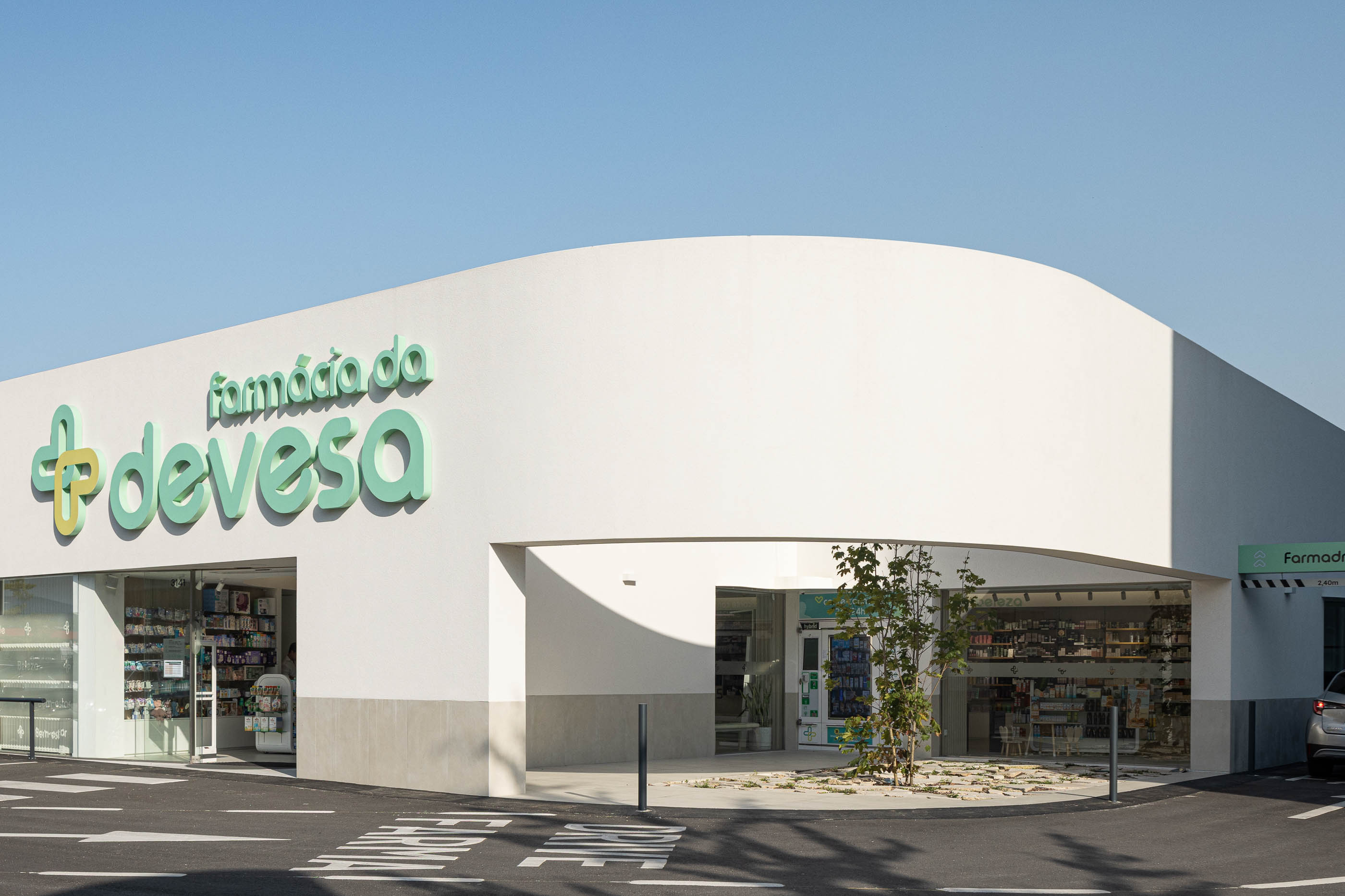 Exterior: Fachada curva branca com entradas distintas, jardim interior e estacionamento. — Farmácia da Devesa arquitetura Tsou Arquitectos em Vila Nova de Famalicão